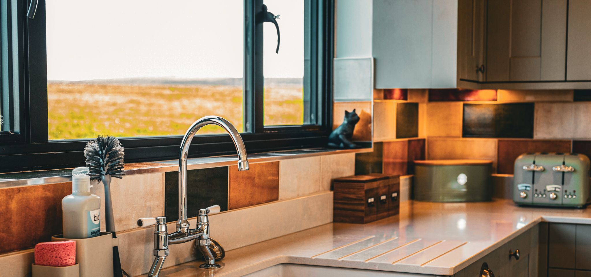 Modern kitchen sink with chrome tap beneath a window, with tiled splashback and countertop appliances