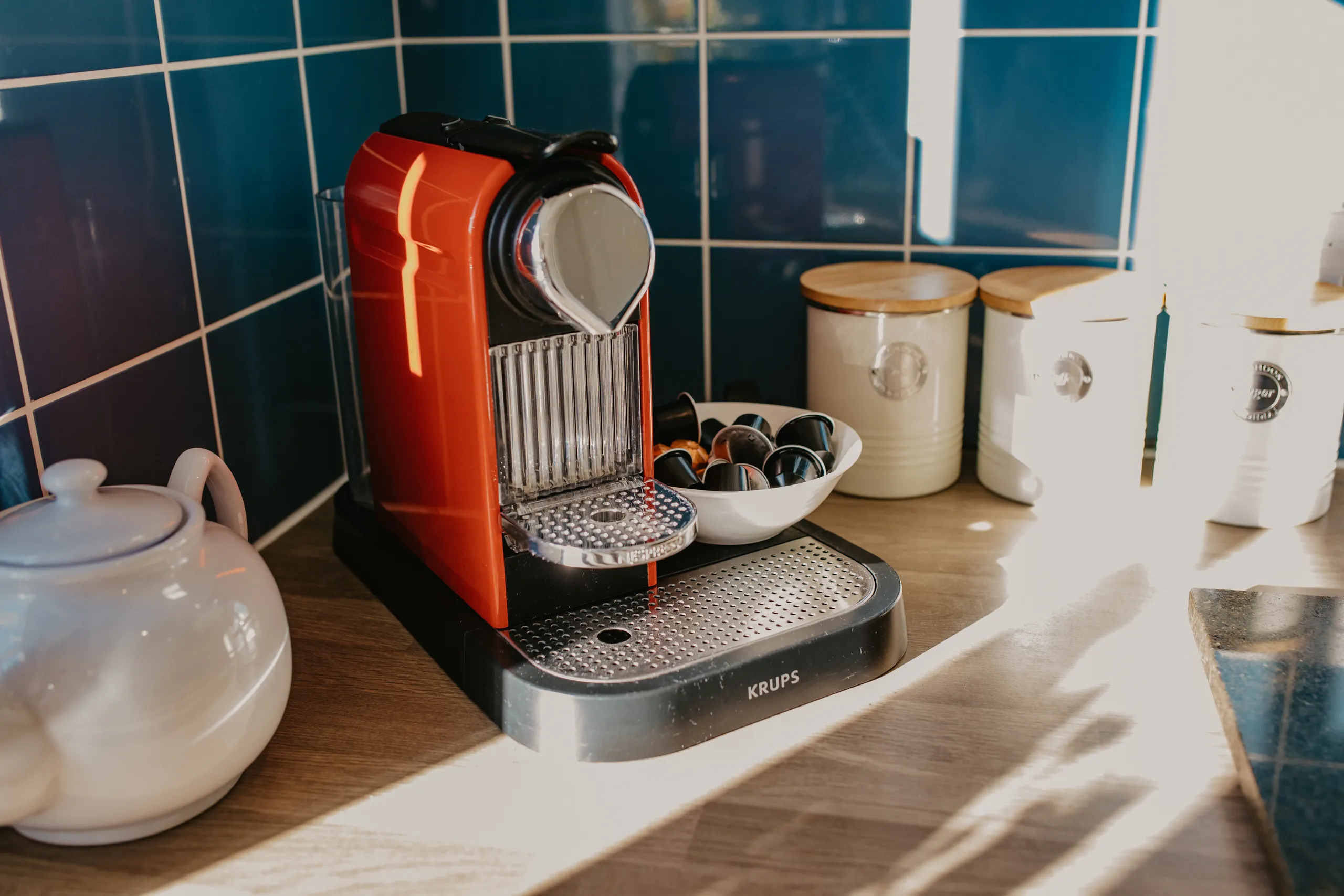 Red coffee pod machine on a wooden kitchen worktop beside cream storage jars and a bowl of capsules