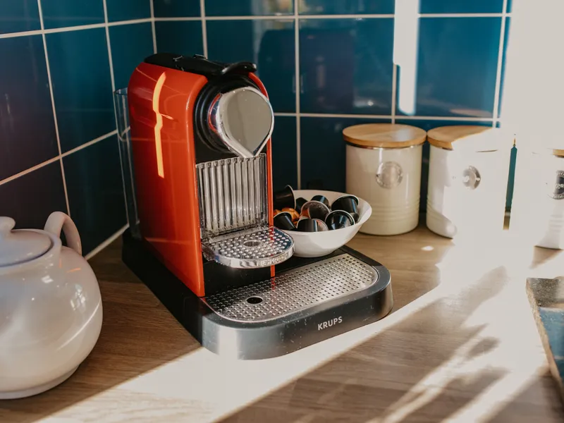 Red coffee pod machine on a wooden kitchen worktop beside cream storage jars and a bowl of capsules