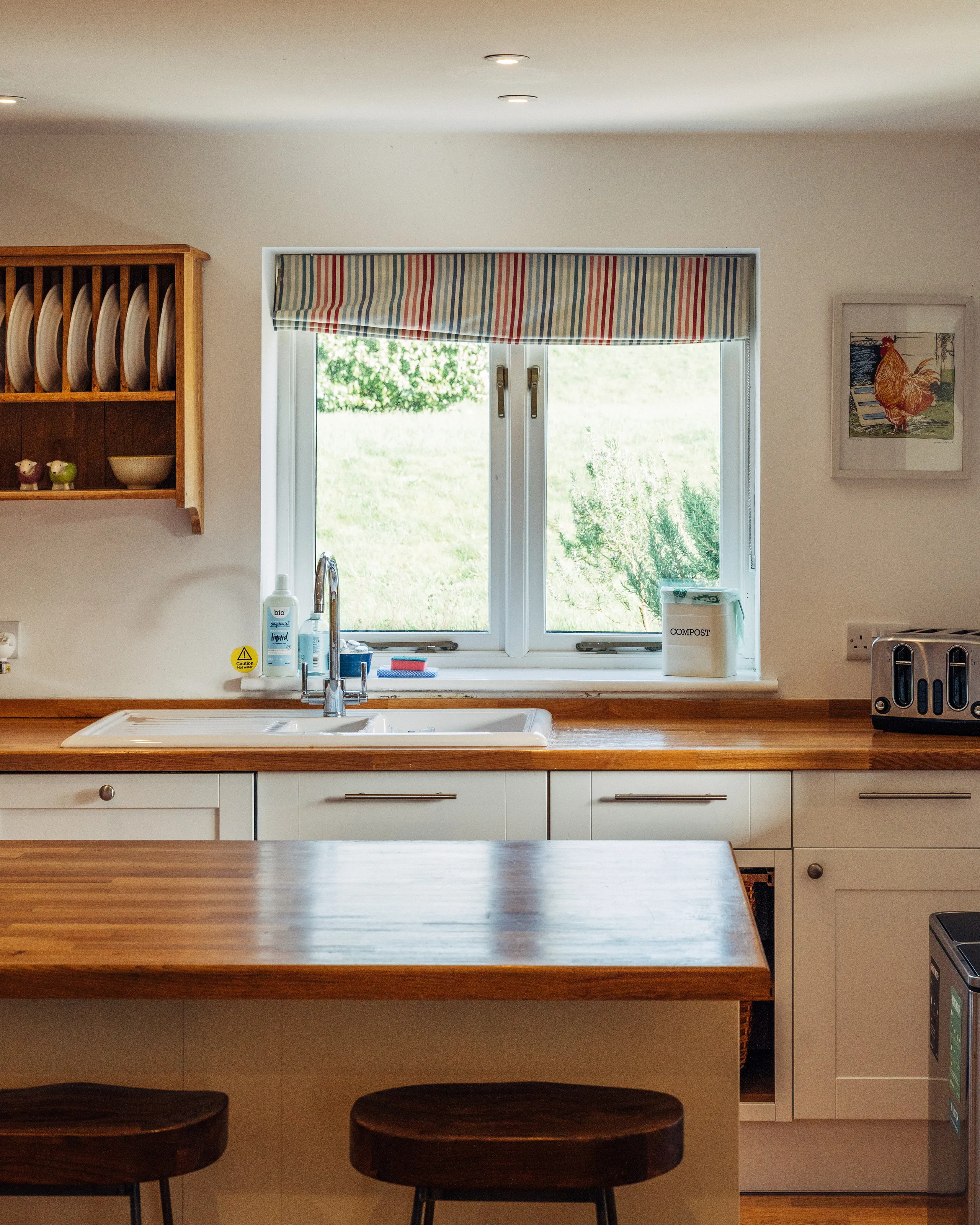 Bright farmhouse kitchen with wooden worktops, a central island, Belfast sink and striped Roman blind over the window.