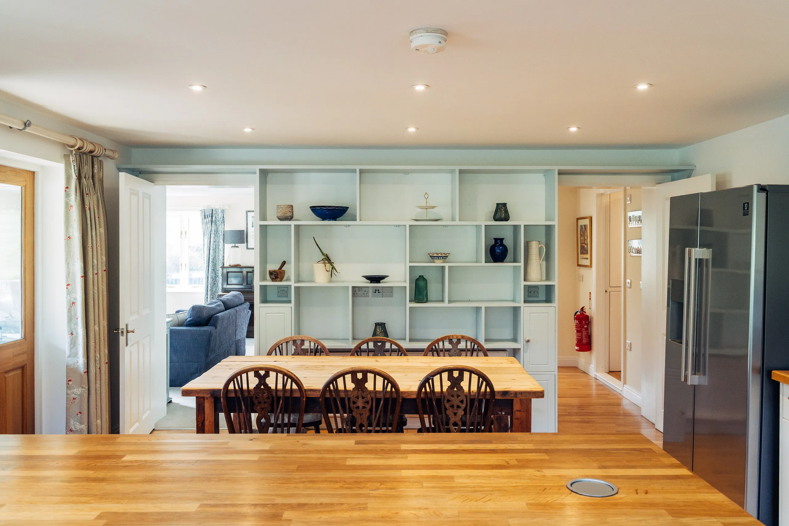 Bright farmhouse-style kitchen diner with a large wooden table, built-in shelving and a stainless steel American-style fridge.