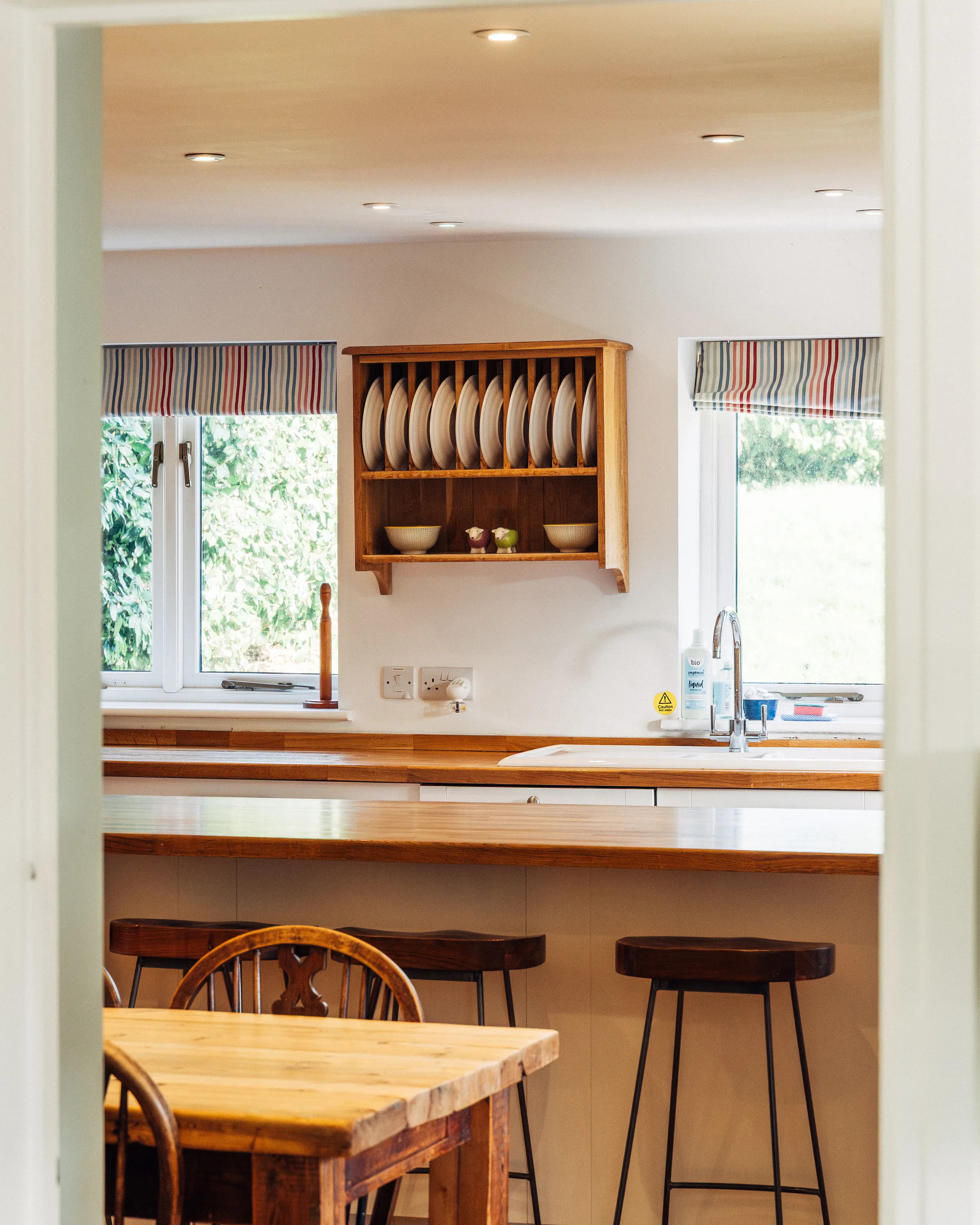 Bright farmhouse kitchen with wooden worktops, striped Roman blinds, a Belfast-style sink and bar stools