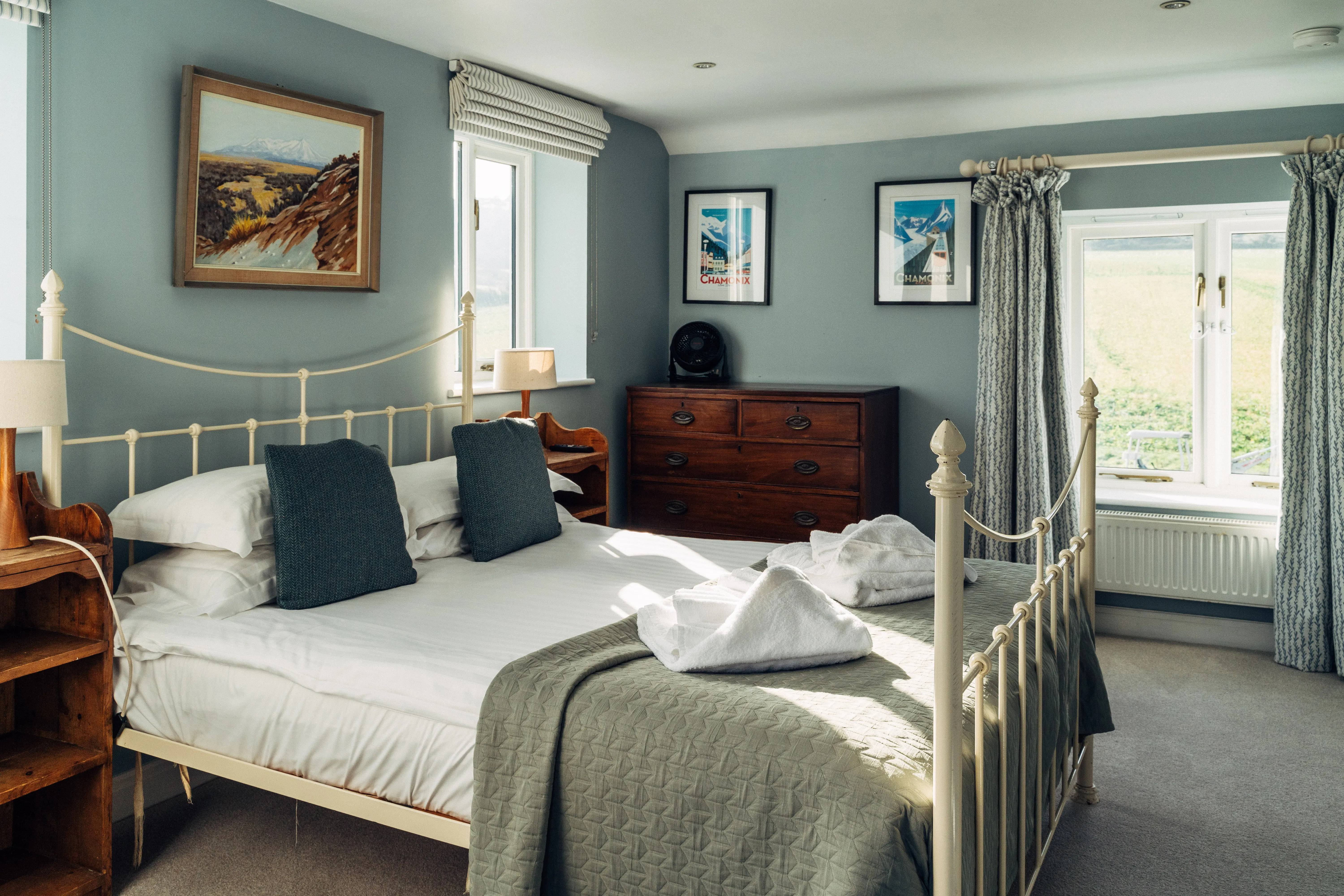 Bright, cosy bedroom with a cream metal bed, blue-grey walls, wooden furniture and framed mountain prints.