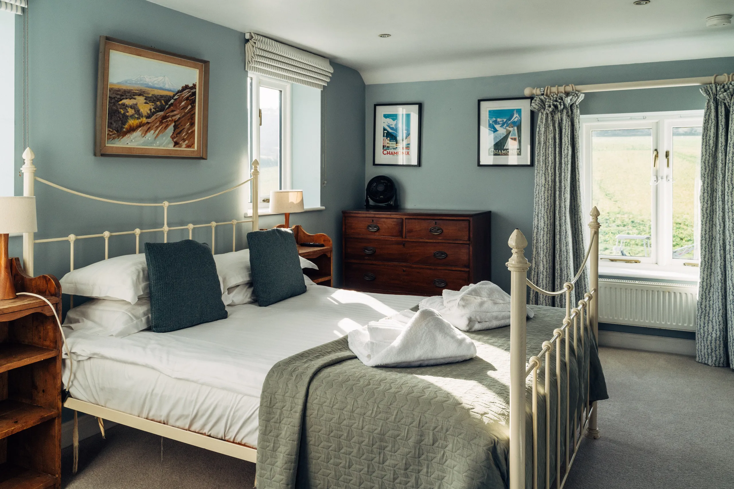 Bright, cosy bedroom with a cream metal bed, blue-grey walls, wooden furniture and framed mountain prints.