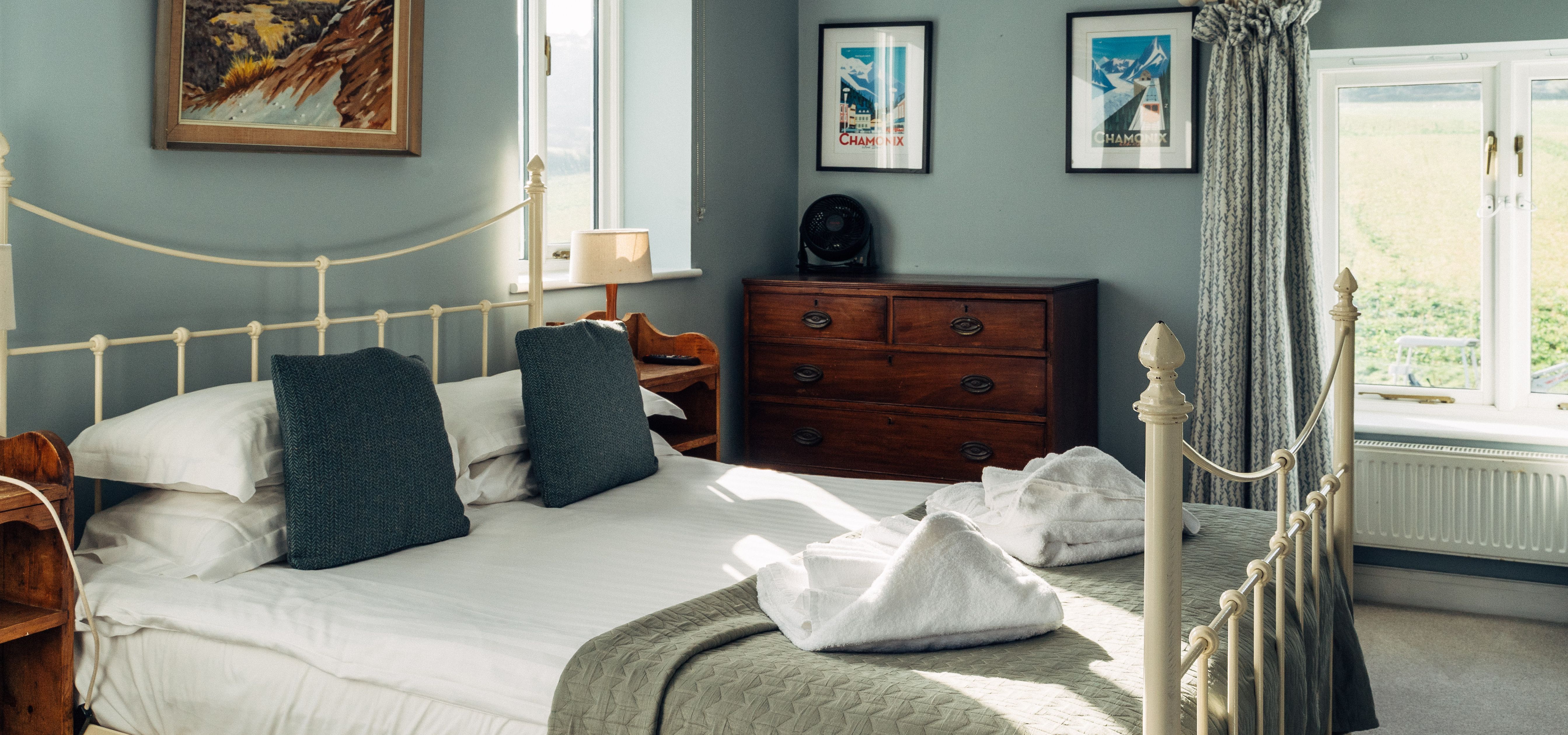 Bright, cosy bedroom with a cream metal bed, blue-grey walls, wooden furniture and framed mountain prints.