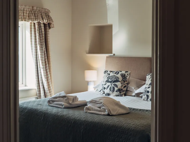 Cosy bedroom with a neatly made double bed, patterned cushions, folded towels, and soft light from a window and bedside lamp.