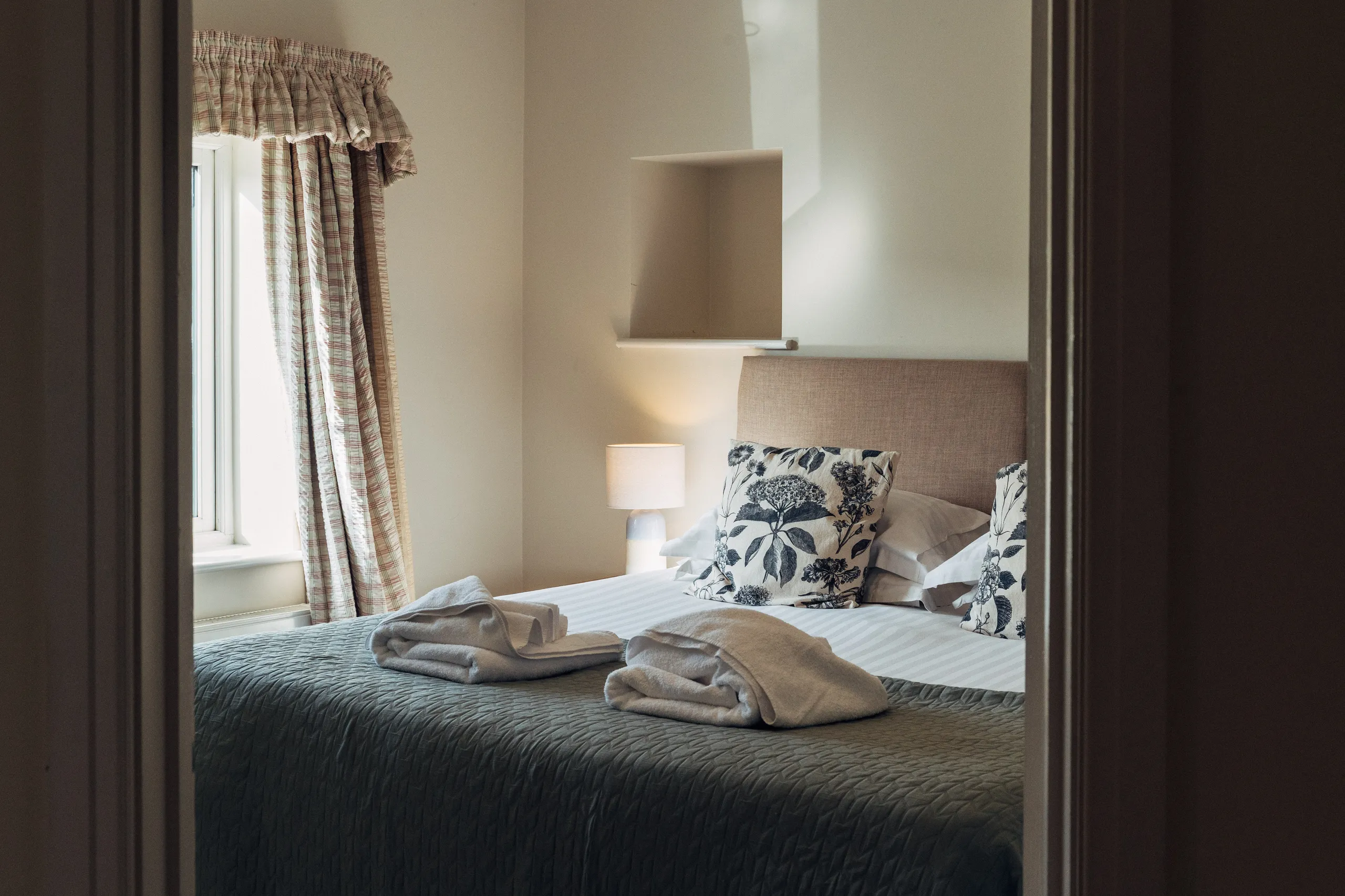 Cosy bedroom with a neatly made double bed, patterned cushions, folded towels, and soft light from a window and bedside lamp.