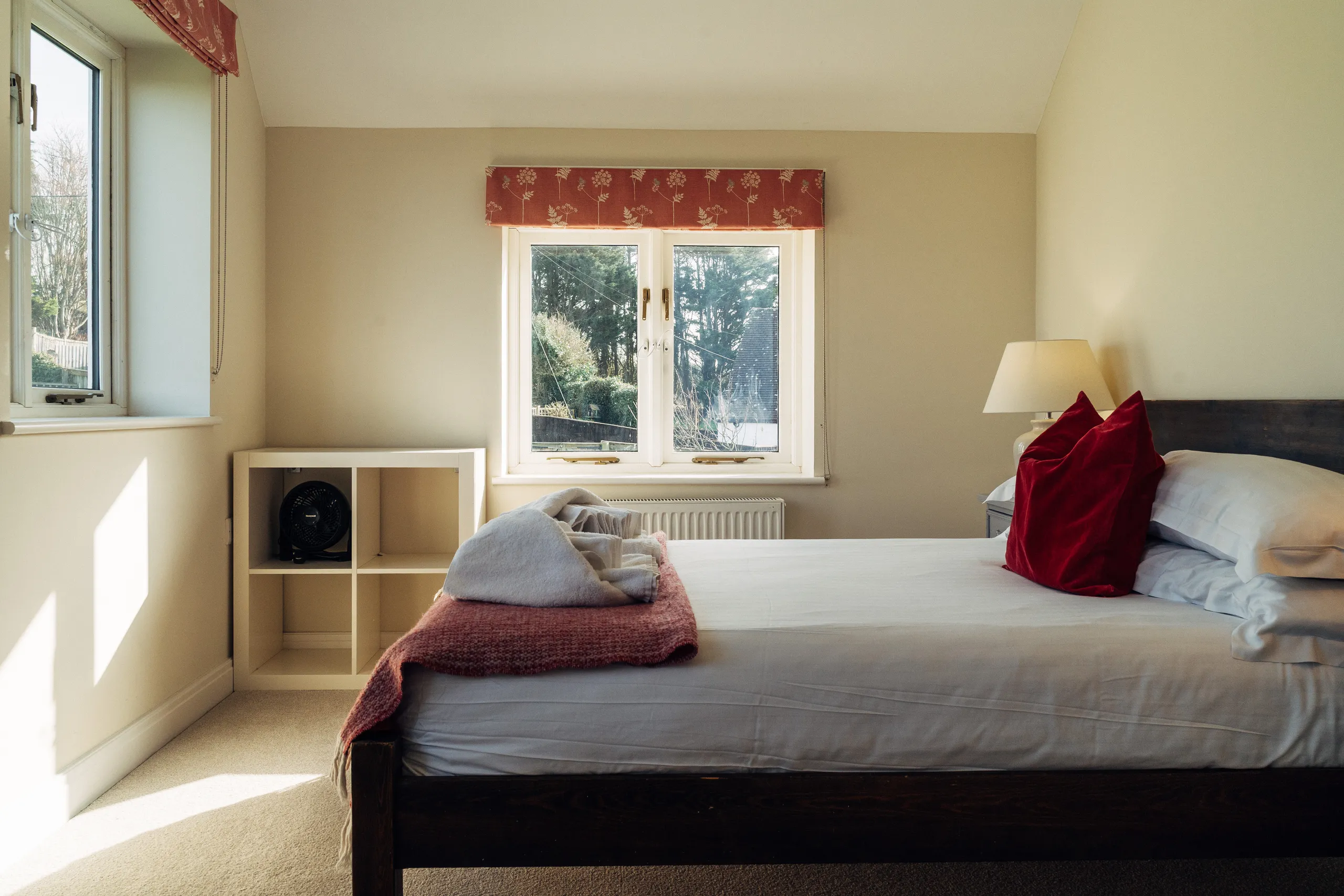 Bright bedroom with a wooden bed, white bedding, red cushions and sunlit windows