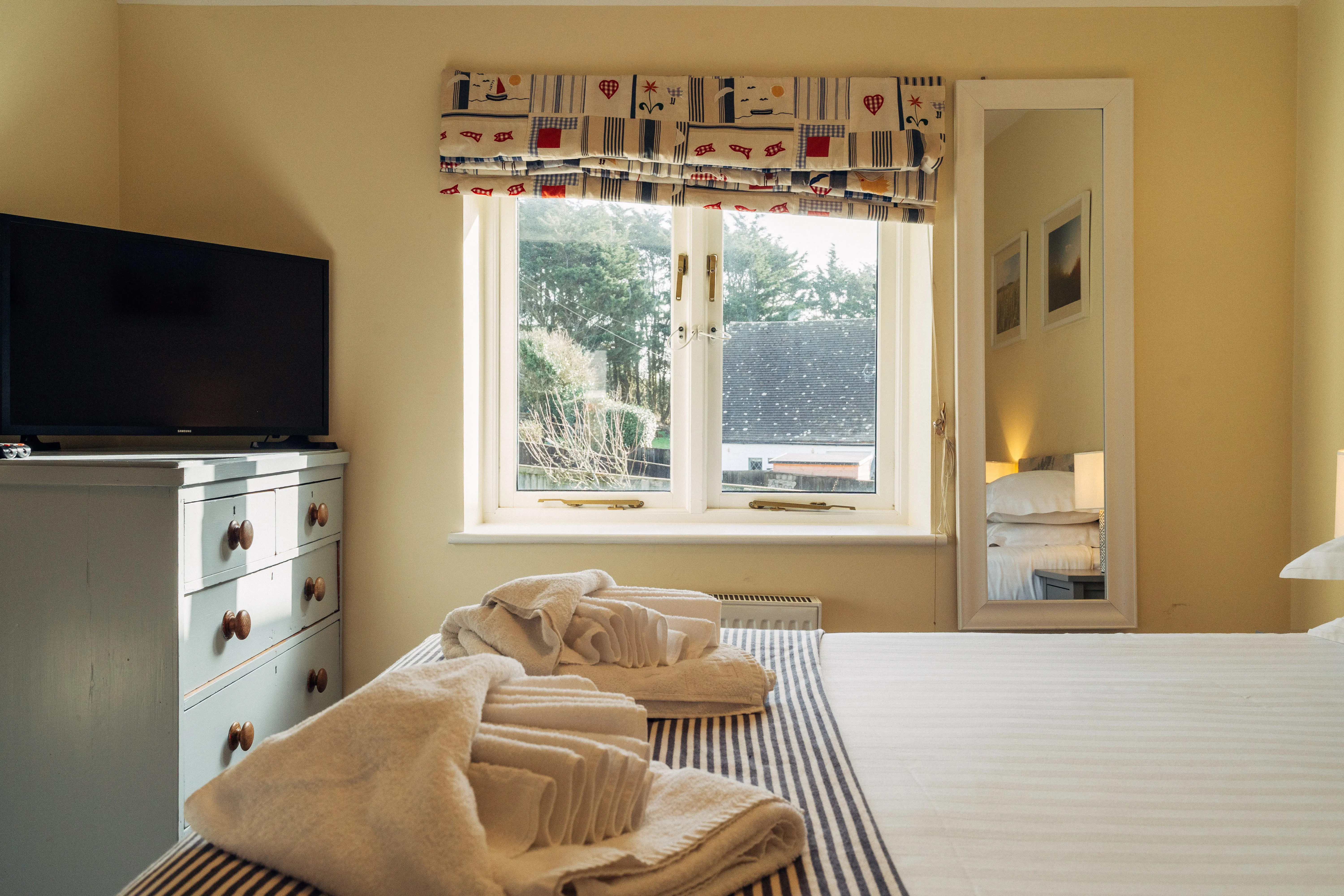 Cosy bedroom with neatly folded towels on the bed, a window with patterned Roman blind, and a television on a painted chest of drawers.
