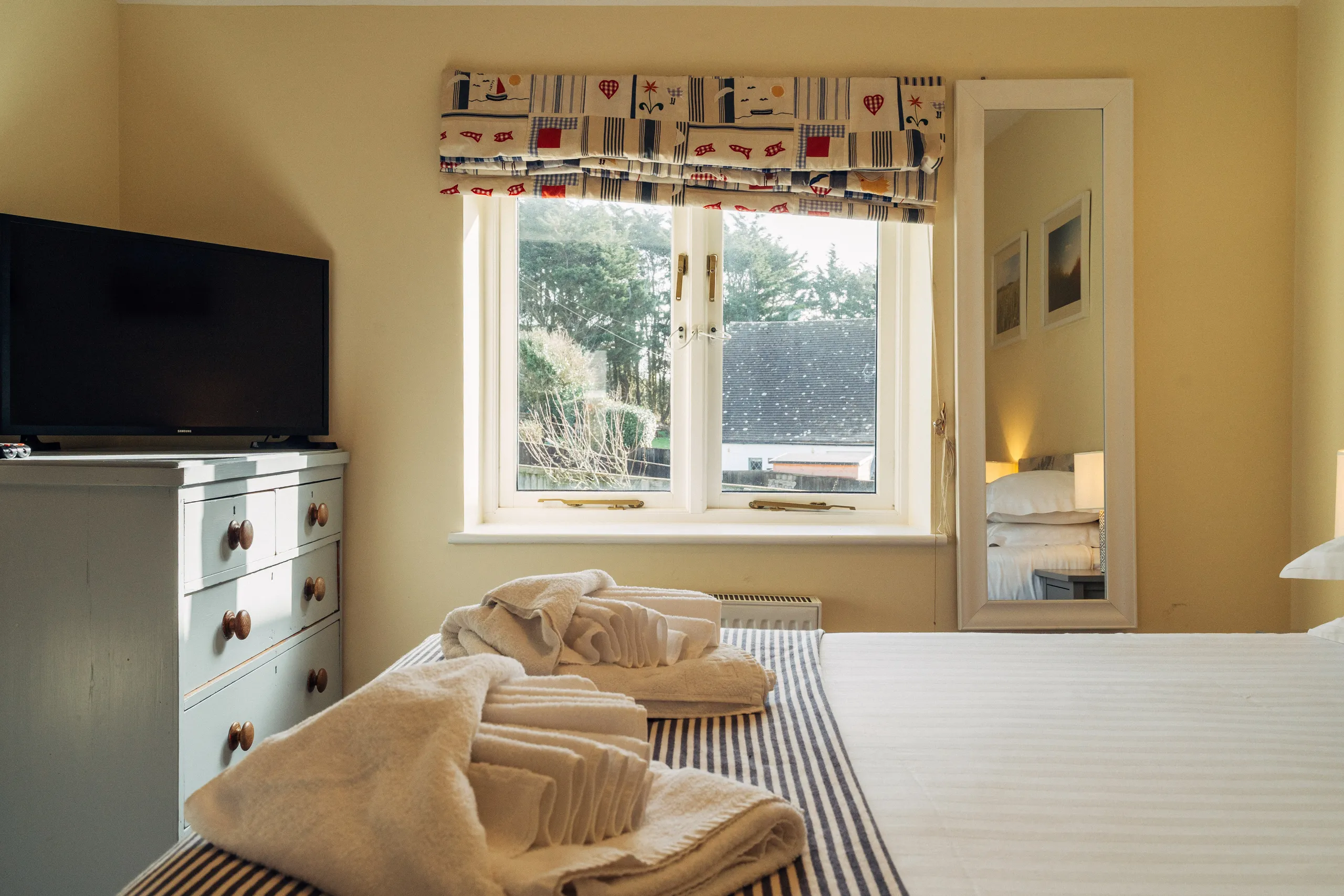 Cosy bedroom with neatly folded towels on the bed, a window with patterned Roman blind, and a television on a painted chest of drawers.