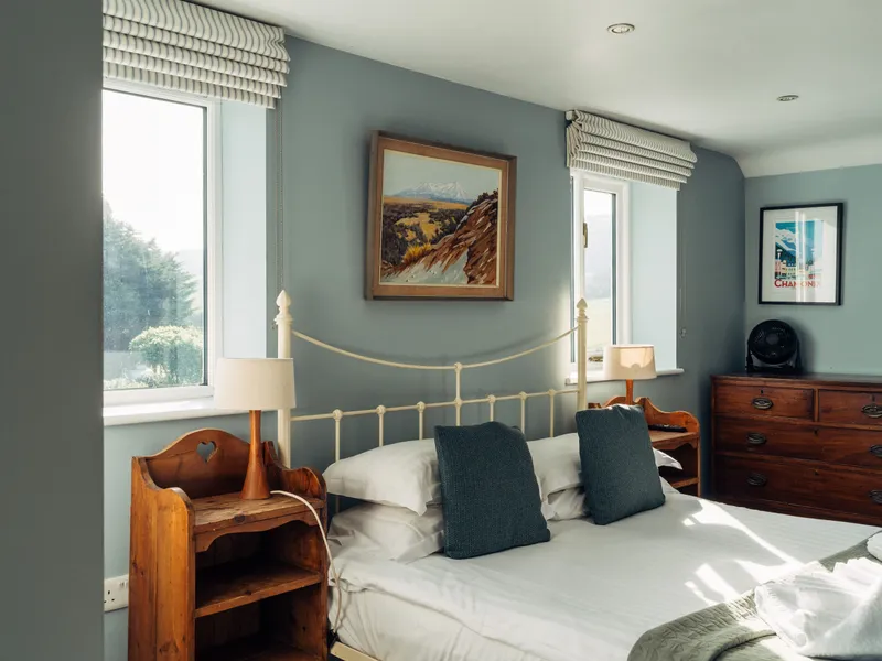 Bright bedroom with a metal bed frame, wooden bedside tables, blue-grey walls and large windows letting in natural light.