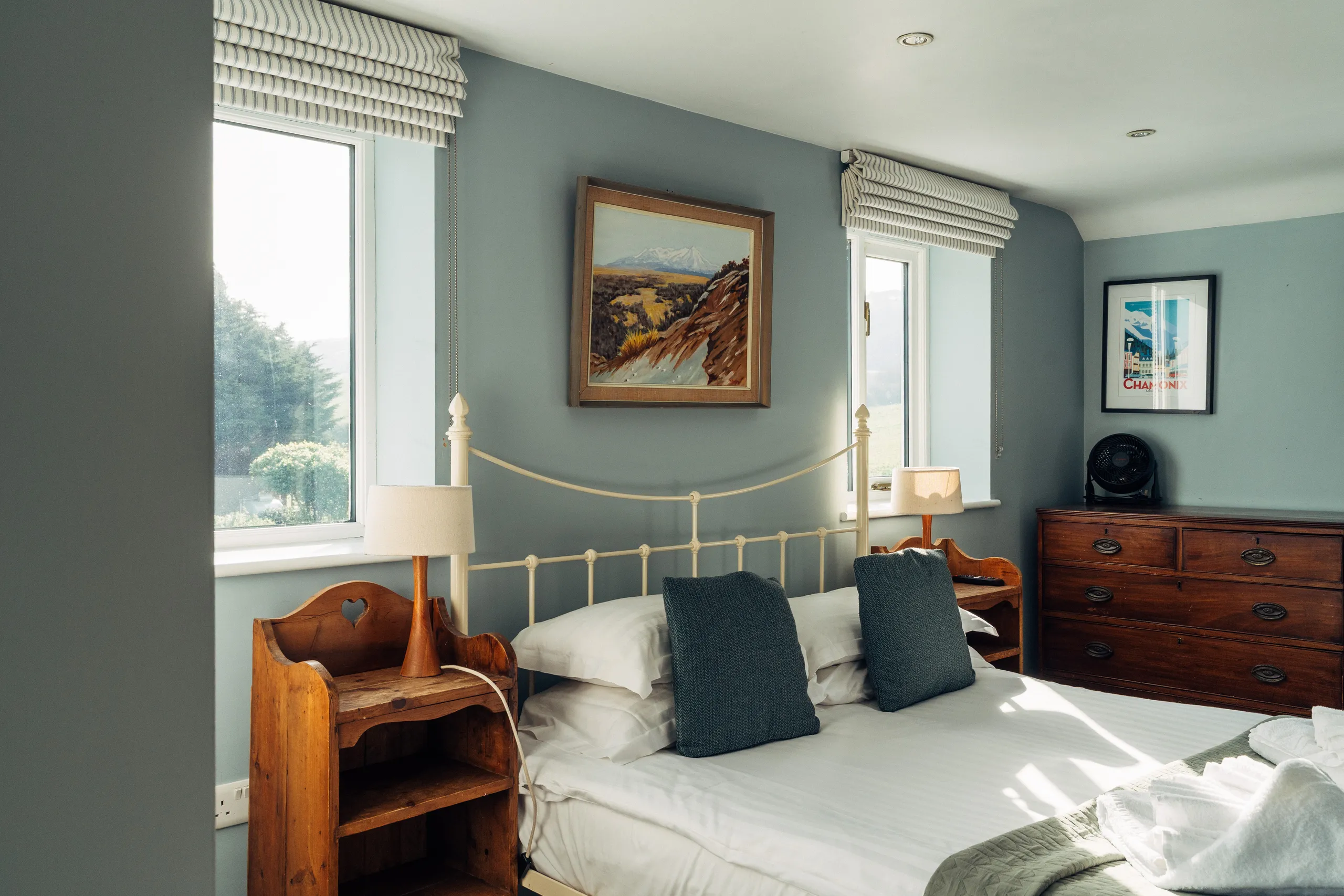 Bright bedroom with a metal bed frame, wooden bedside tables, blue-grey walls and large windows letting in natural light.