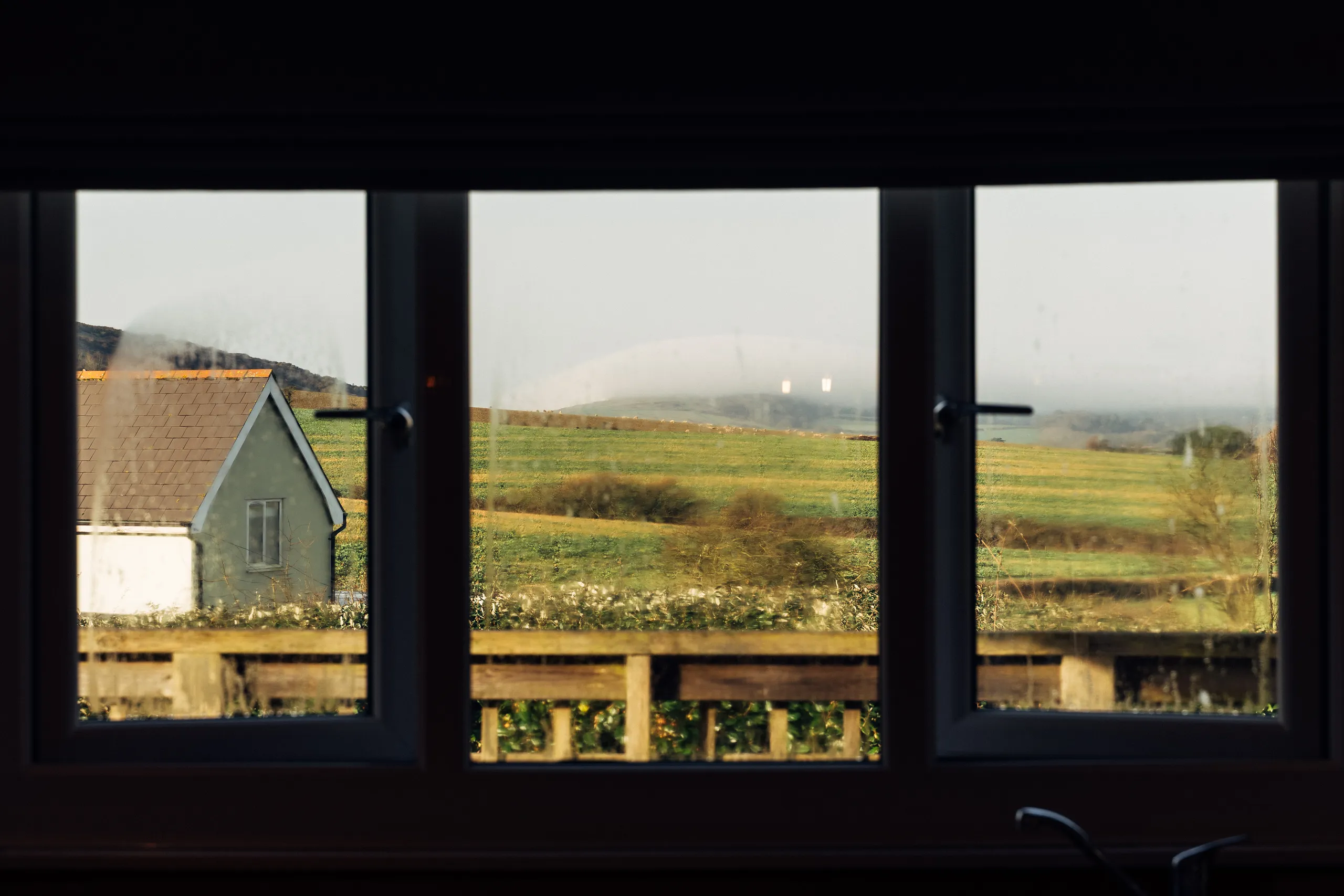 View through a cottage window across green winter fields and a neighbouring house in the countryside