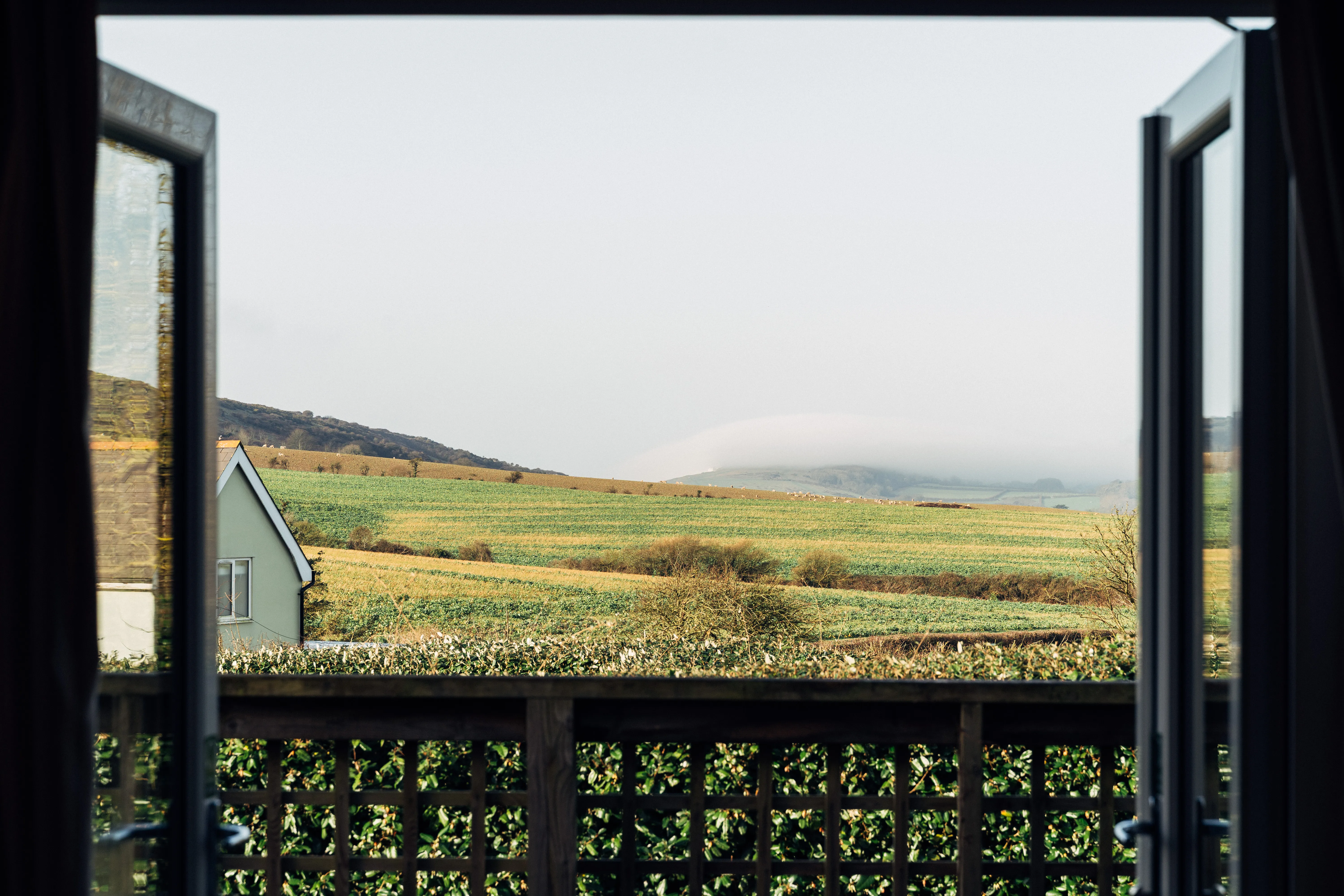 View through open patio doors across rolling green and golden winter fields with mist-covered hills in the distance