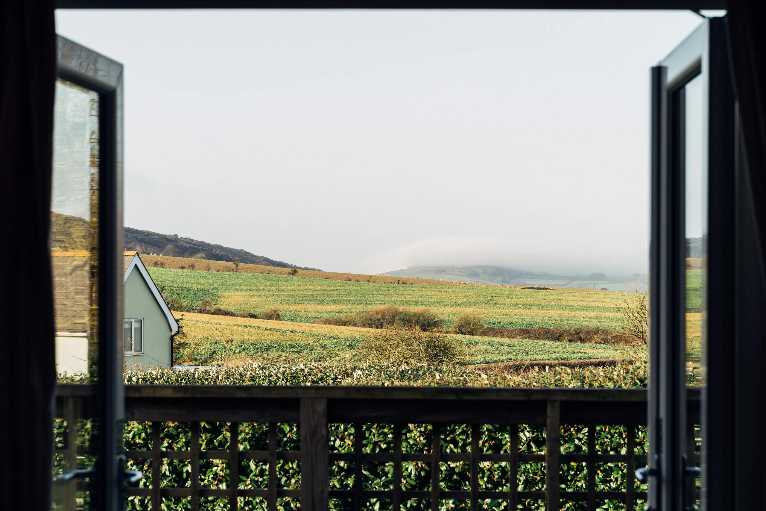 View through open patio doors across rolling green and golden winter fields with mist-covered hills in the distance