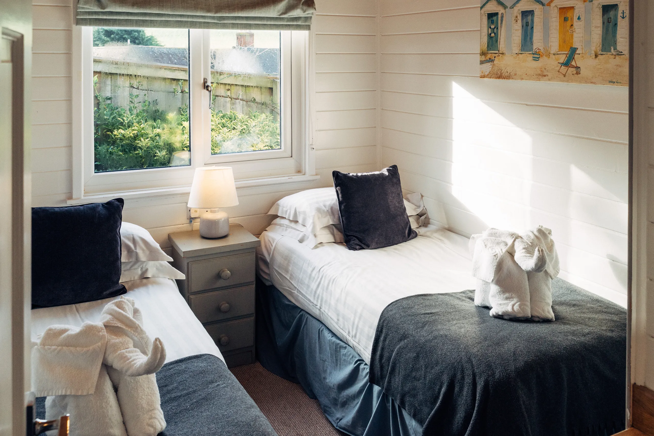 Twin bedroom with two single beds, a bedside table and lamp, dark grey throws, and towel animals in a bright coastal-style interior