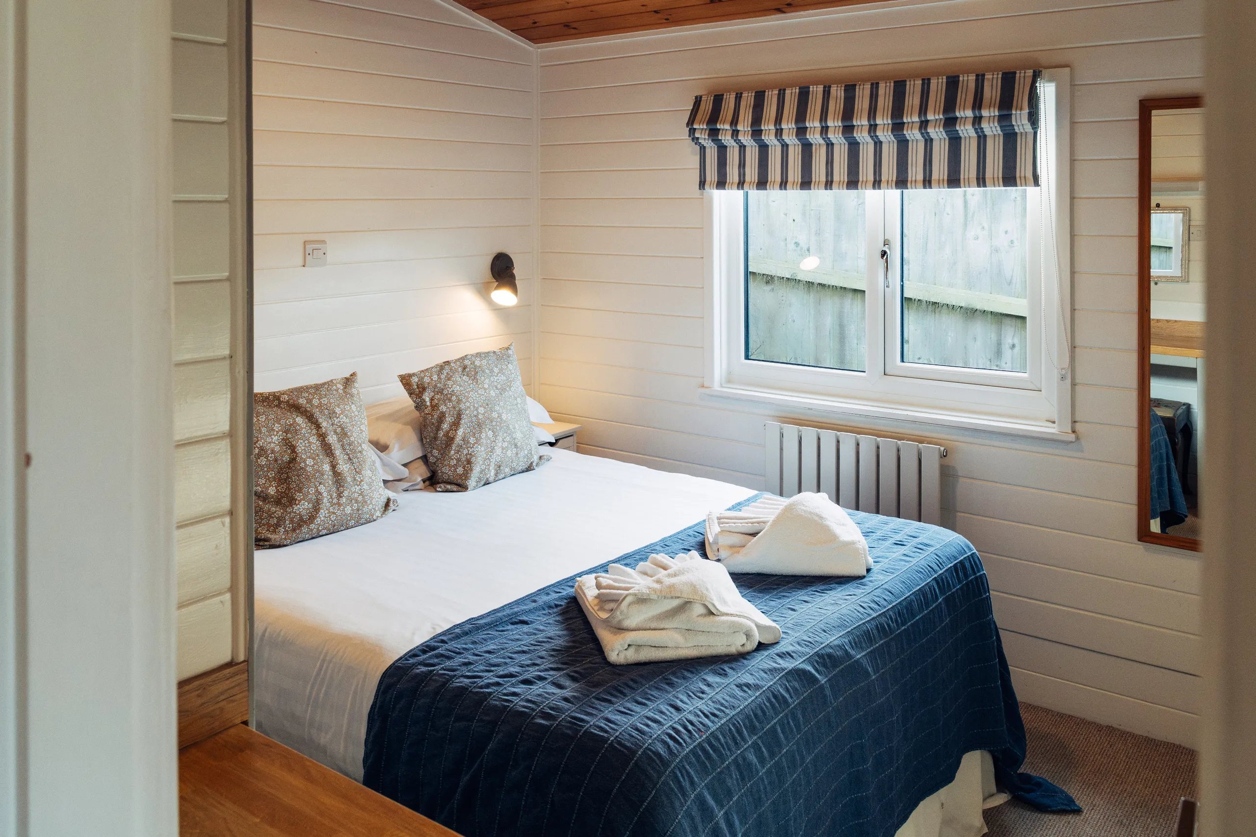 Cosy bedroom with a double bed, blue bedspread, folded towels, striped blind and wooden panelled walls