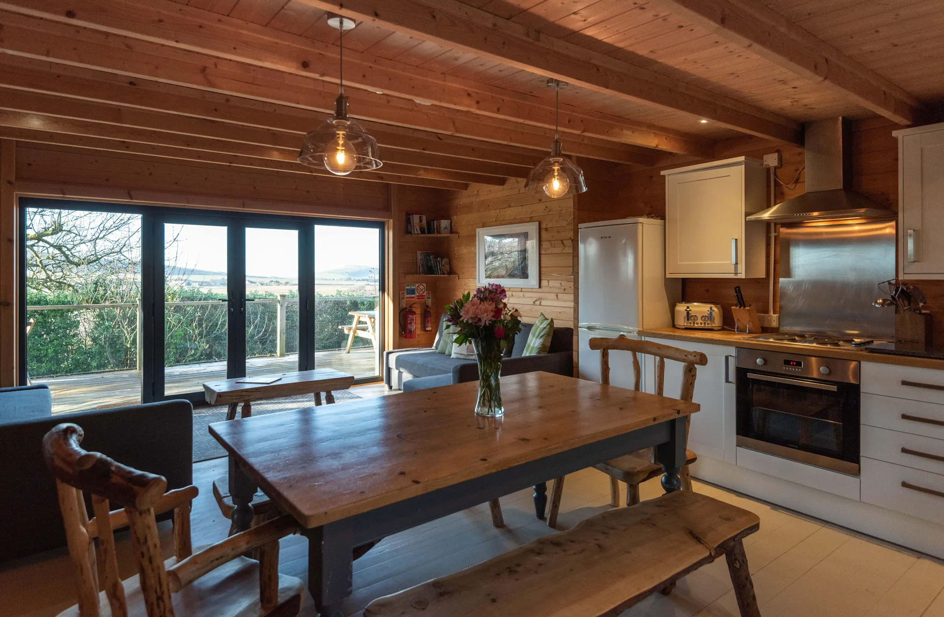 Cozy wooden cabin kitchen and dining area with rustic furniture, large windows, and countryside view