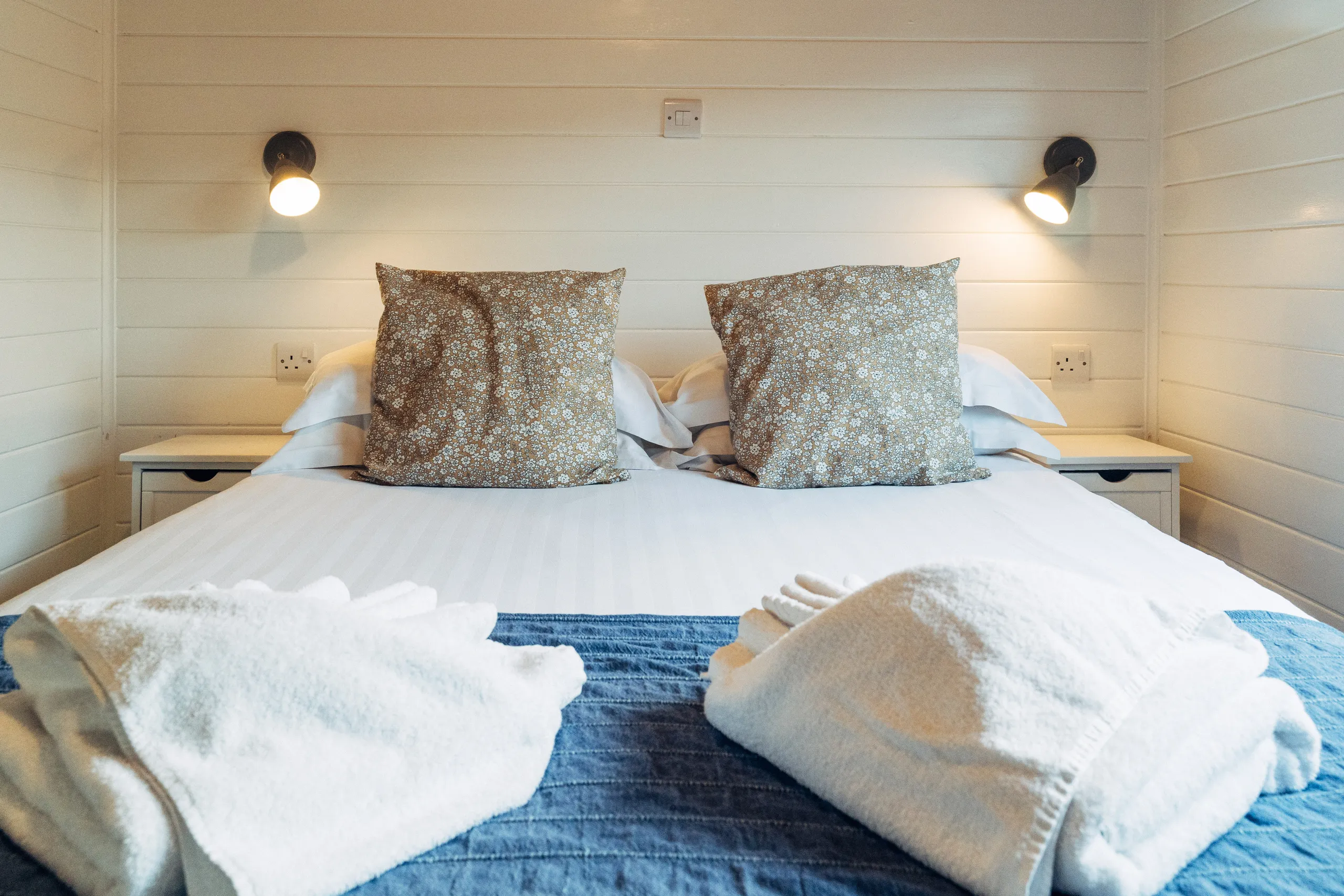 Double bed with white linens, floral cushions and folded towels in a cosy wood-panelled bedroom