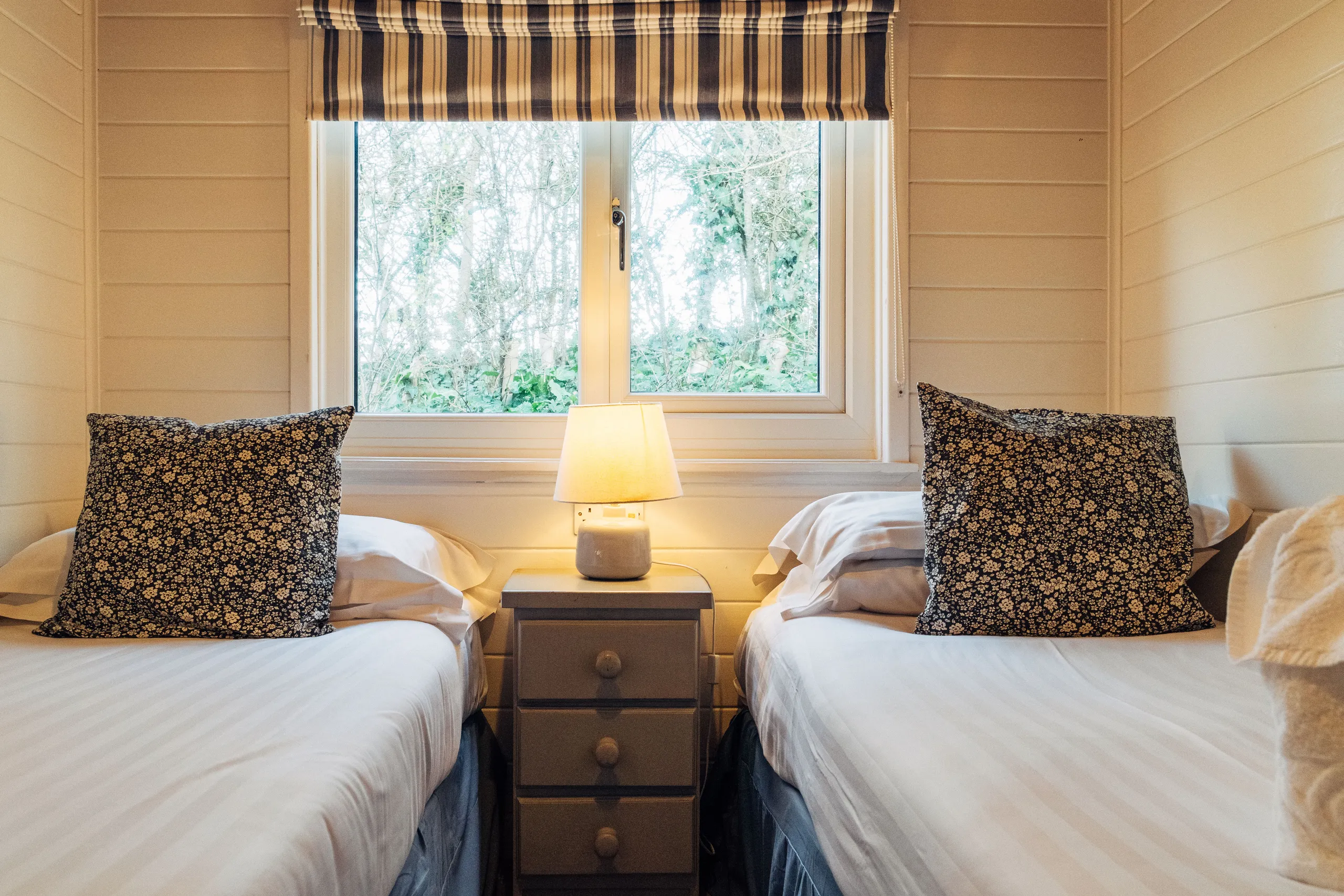 Twin bedroom with two single beds, floral cushions, a bedside lamp and a window with striped Roman blind
