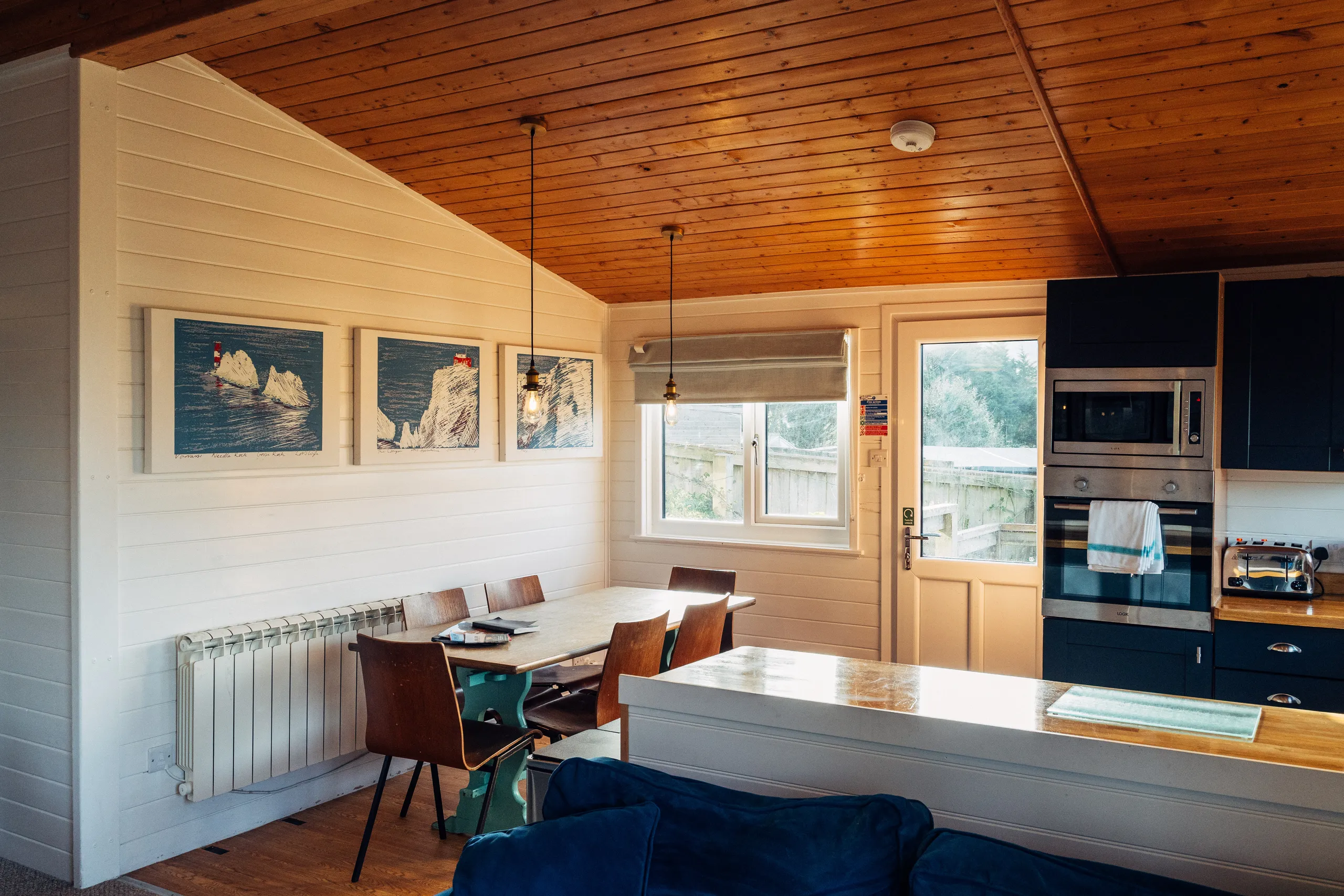 Open-plan lodge kitchen and dining area with wooden ceiling, framed coastal artwork and navy cabinets