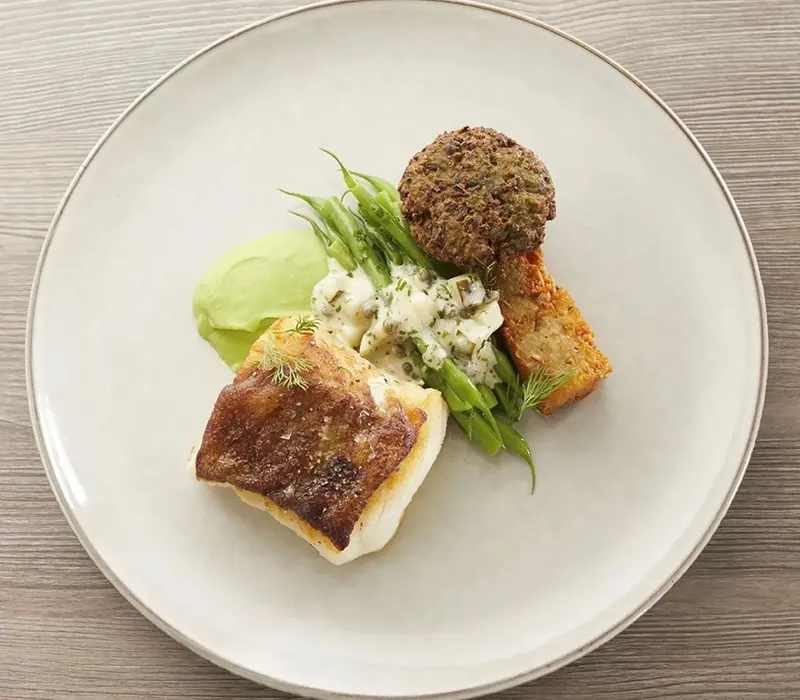 Plated fish dish with crispy-skinned fillet, green beans, herb sauce and a croquette on a large ceramic plate