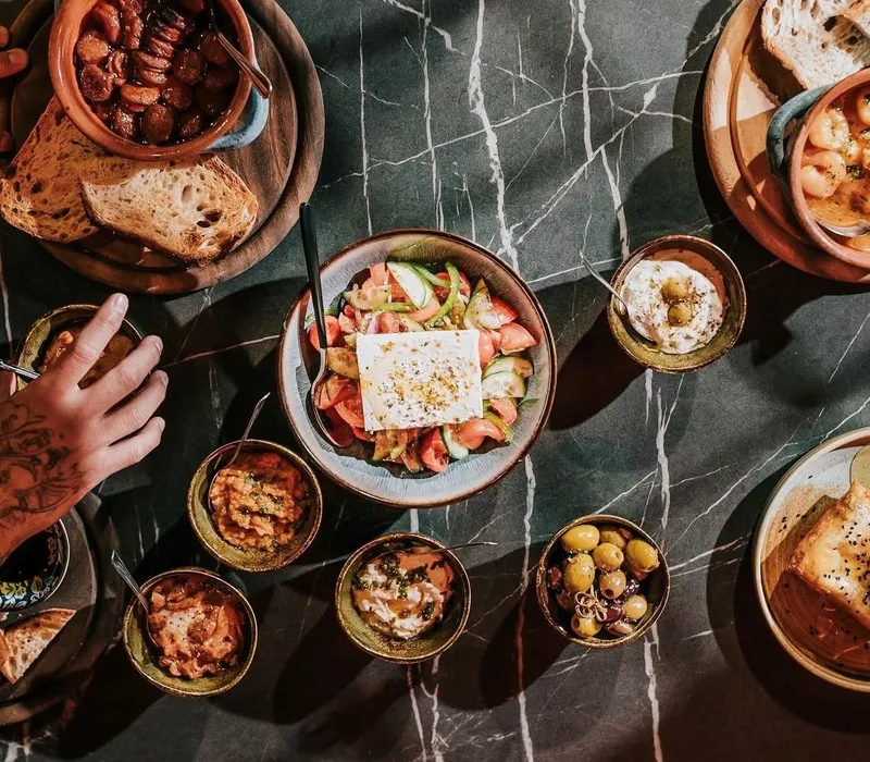 Overhead view of a shared Mediterranean-style meal with bread, salad topped with feta, olives and small dishes on a dark marble table.