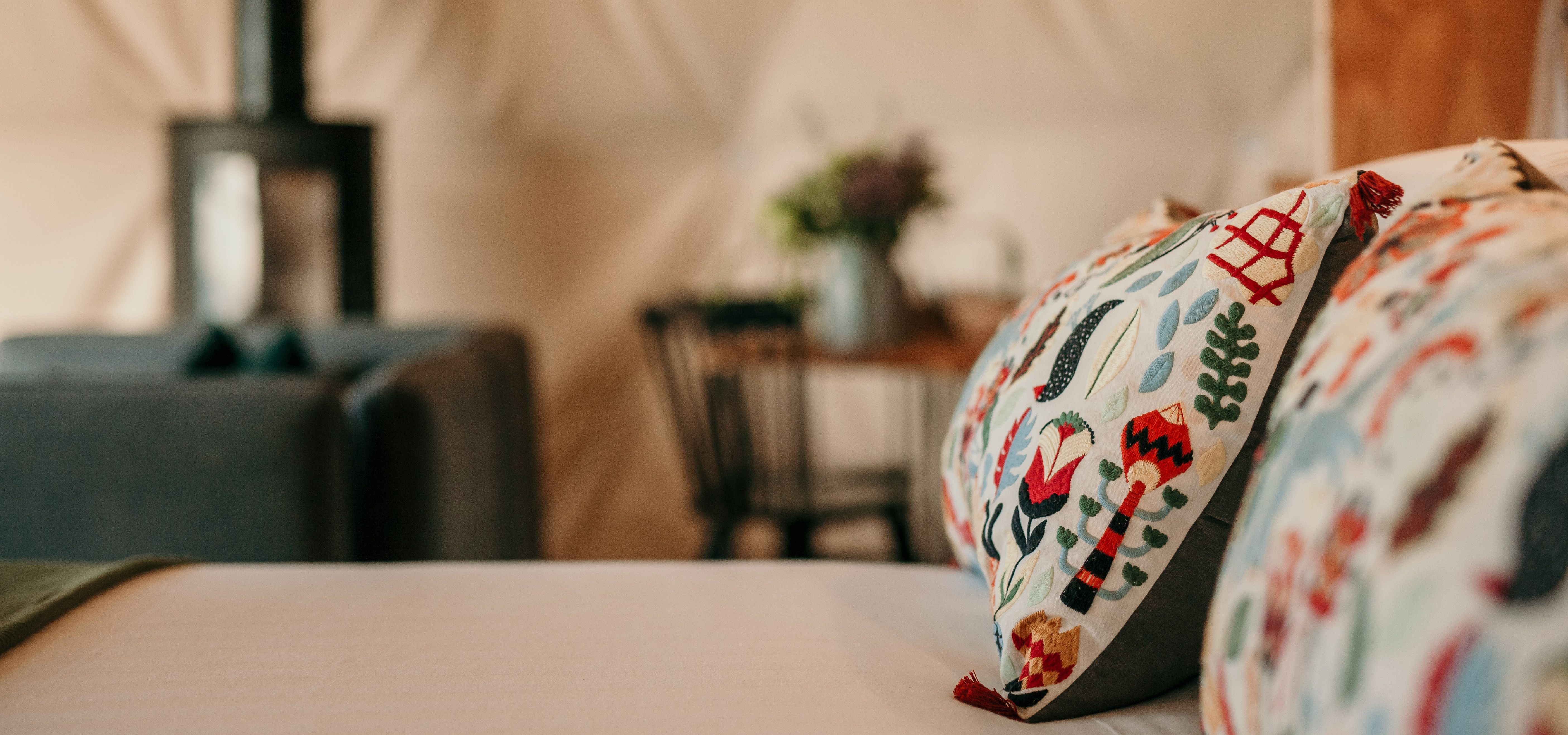 Close-up of colourful embroidered cushions on a bed inside Tom's Eco Lodge dome at Tapnell Farm, with a wood-burning stove in the background.