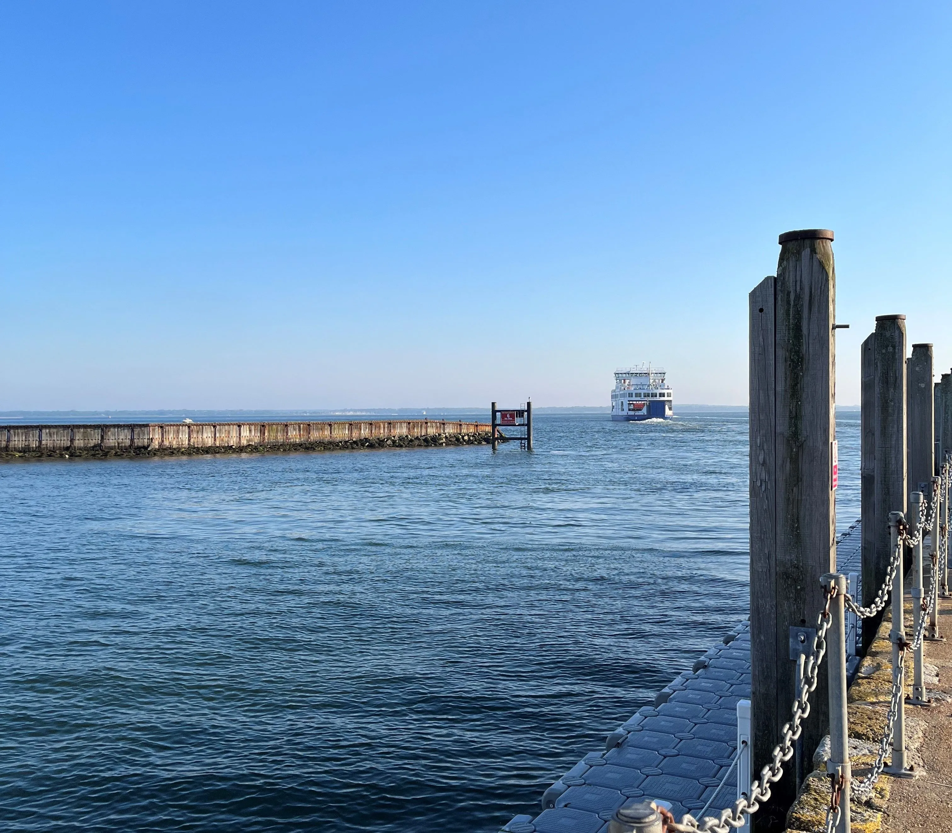 View across Yarmouth Harbour with calm blue water, a distant ferry and wooden mooring posts along the quayside.