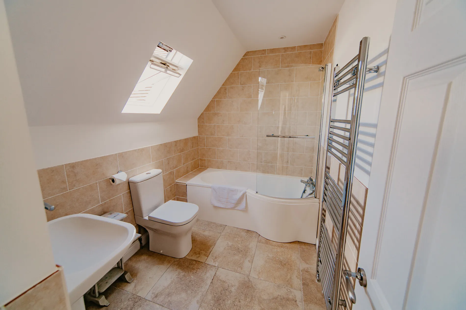 Bright bathroom at Dairyman's Cottage, Tapnell Farm with corner bath, beige tiled walls, toilet and skylight window