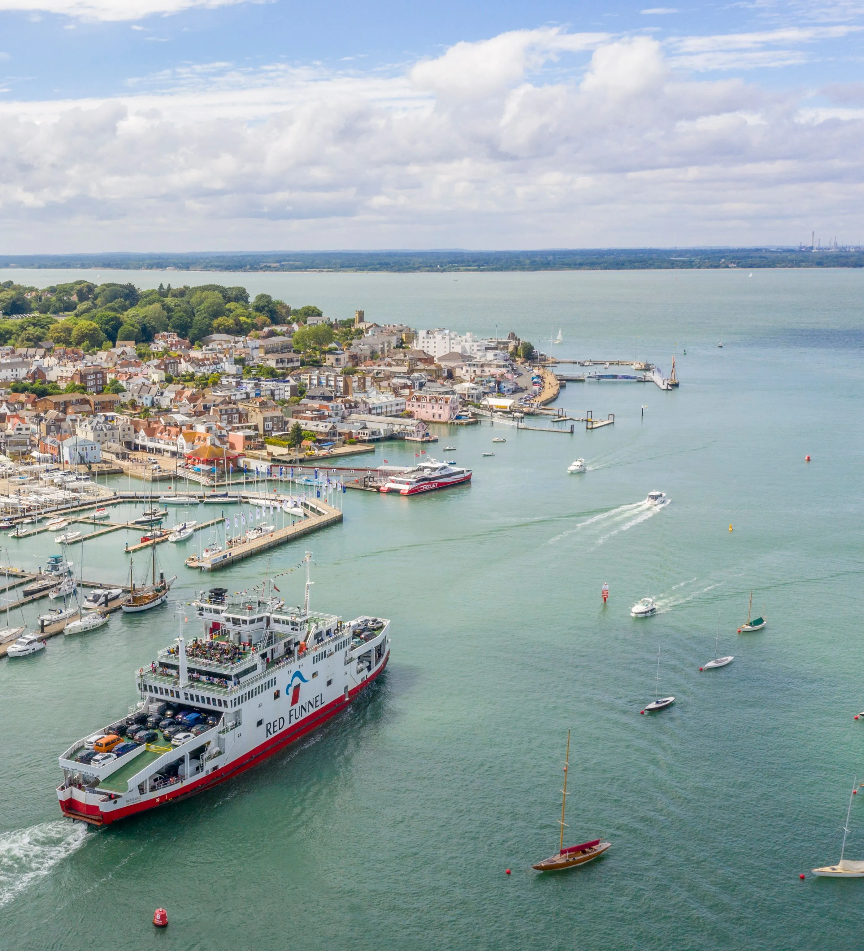 Aerial view of a Red Funnel ferry leaving a marina-lined harbour with yachts and waterfront buildings.