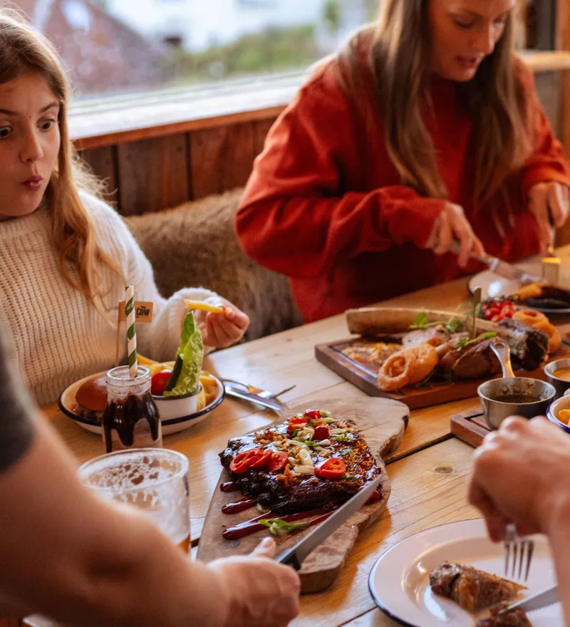 Family enjoying a relaxed meal together at The Cow at Tapnell Farm, with burgers, chips and sharing dishes on a wooden table.