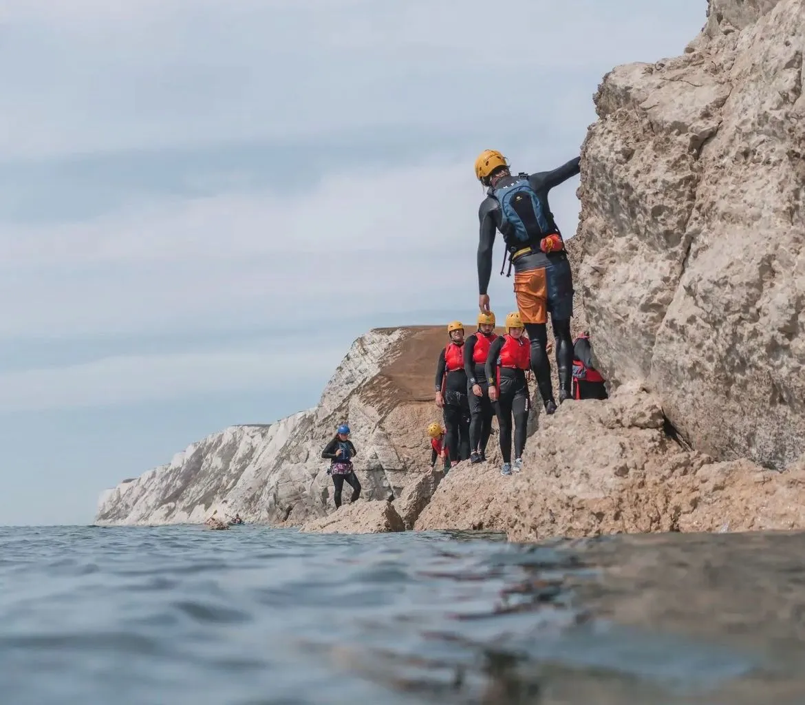 Group coasteering along the rocky shoreline at Freshwater Bay, wearing helmets and buoyancy aids beside white chalk cliffs.