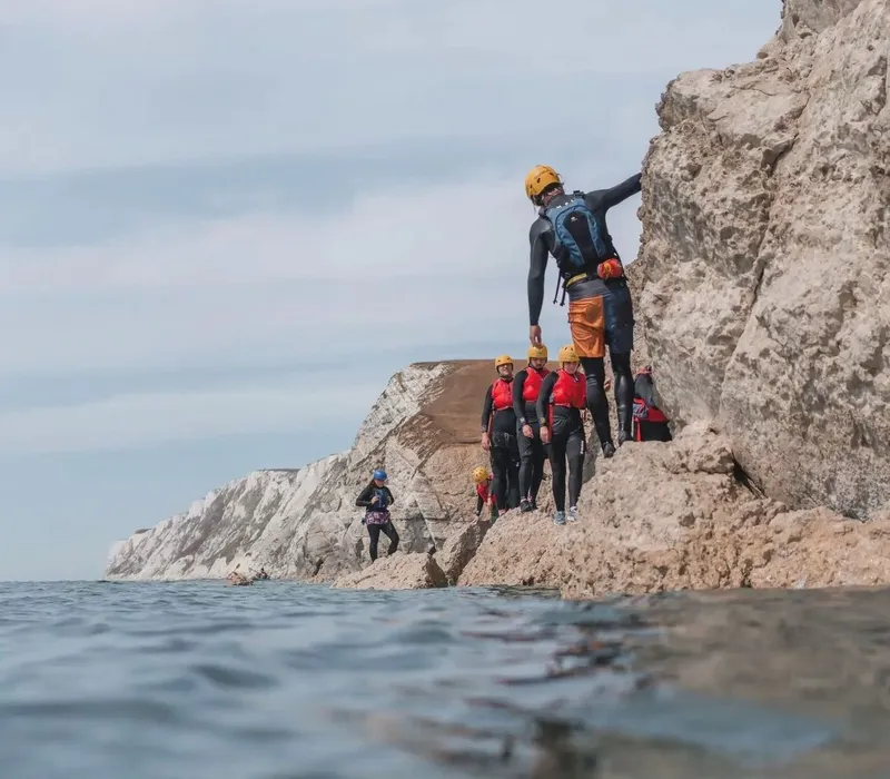 Group coasteering along the rocky shoreline at Freshwater Bay, wearing helmets and buoyancy aids beside white chalk cliffs.