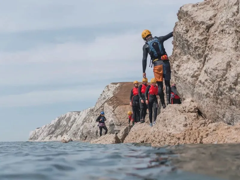 Group coasteering along the rocky shoreline at Freshwater Bay, wearing helmets and buoyancy aids beside white chalk cliffs.