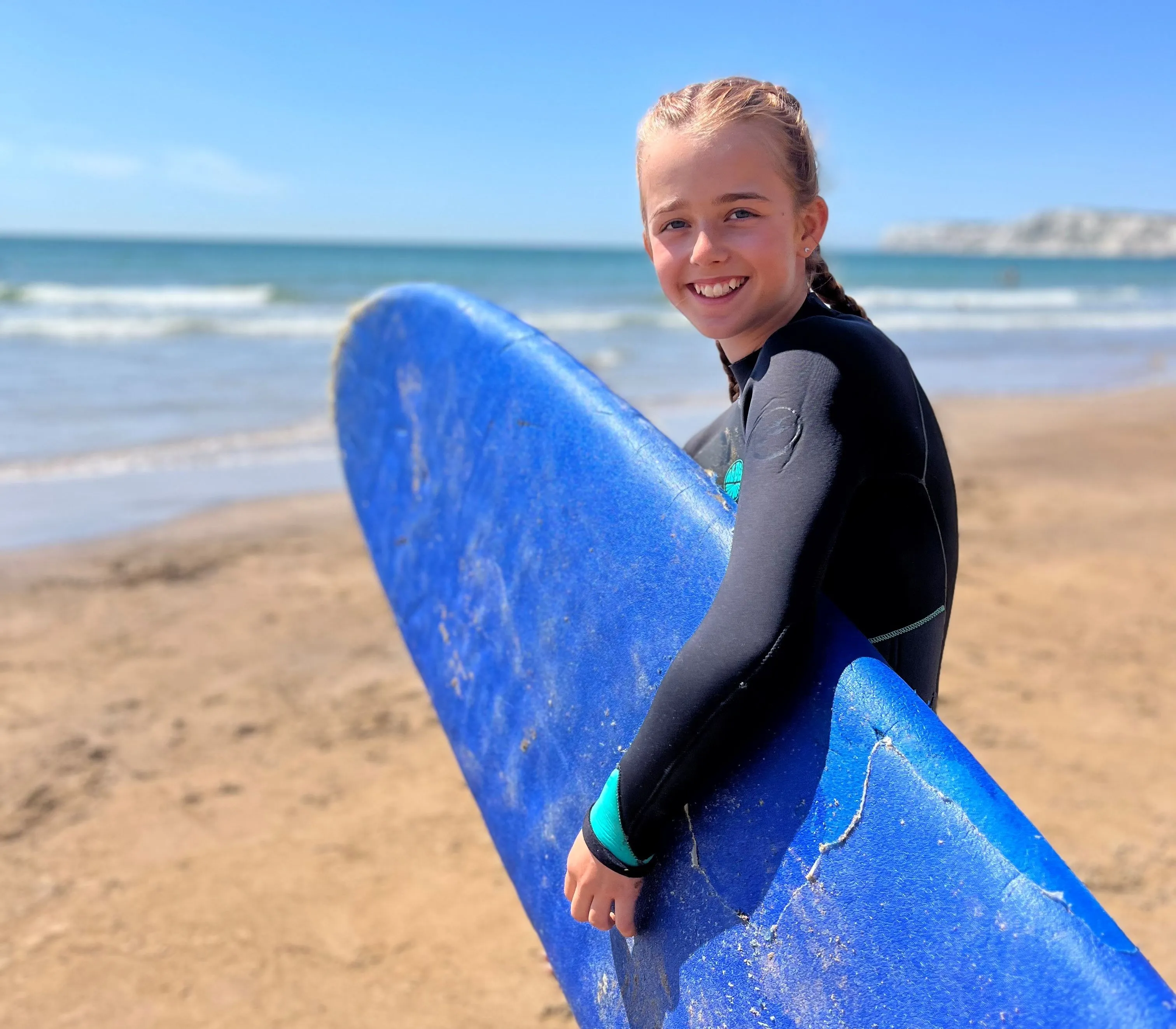 Young girl in a wetsuit holding a blue surfboard on a sandy beach by the sea.