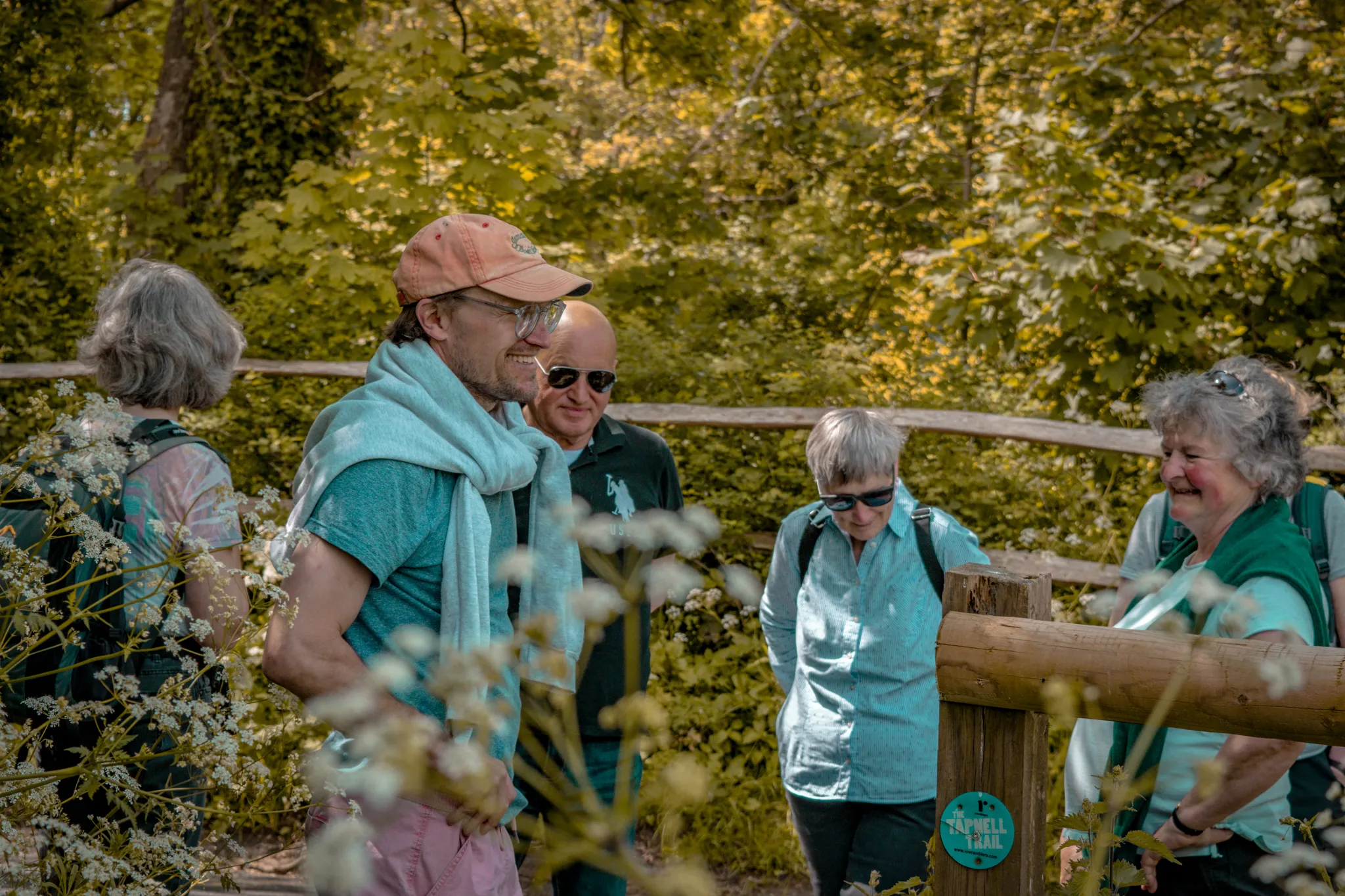 A group of adults chatting beside a wooden gate on the Tapnell Trail in a leafy woodland setting.