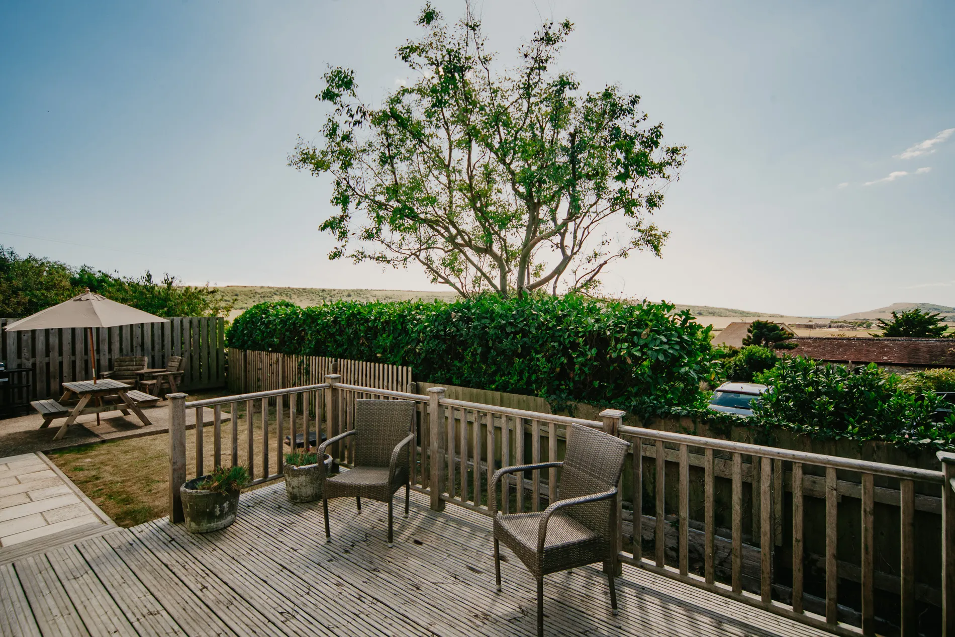 Patio and wooden decking at Dairyman's Cottage, Tapnell Farm, with outdoor chairs, picnic bench and parasol overlooking countryside fields.
