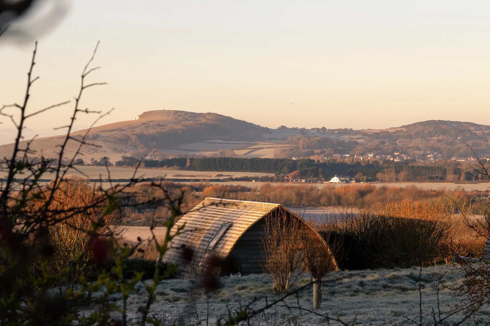A wooden cabin on a frosty winter morning with rolling hills in the background and bare branches in the foreground.
