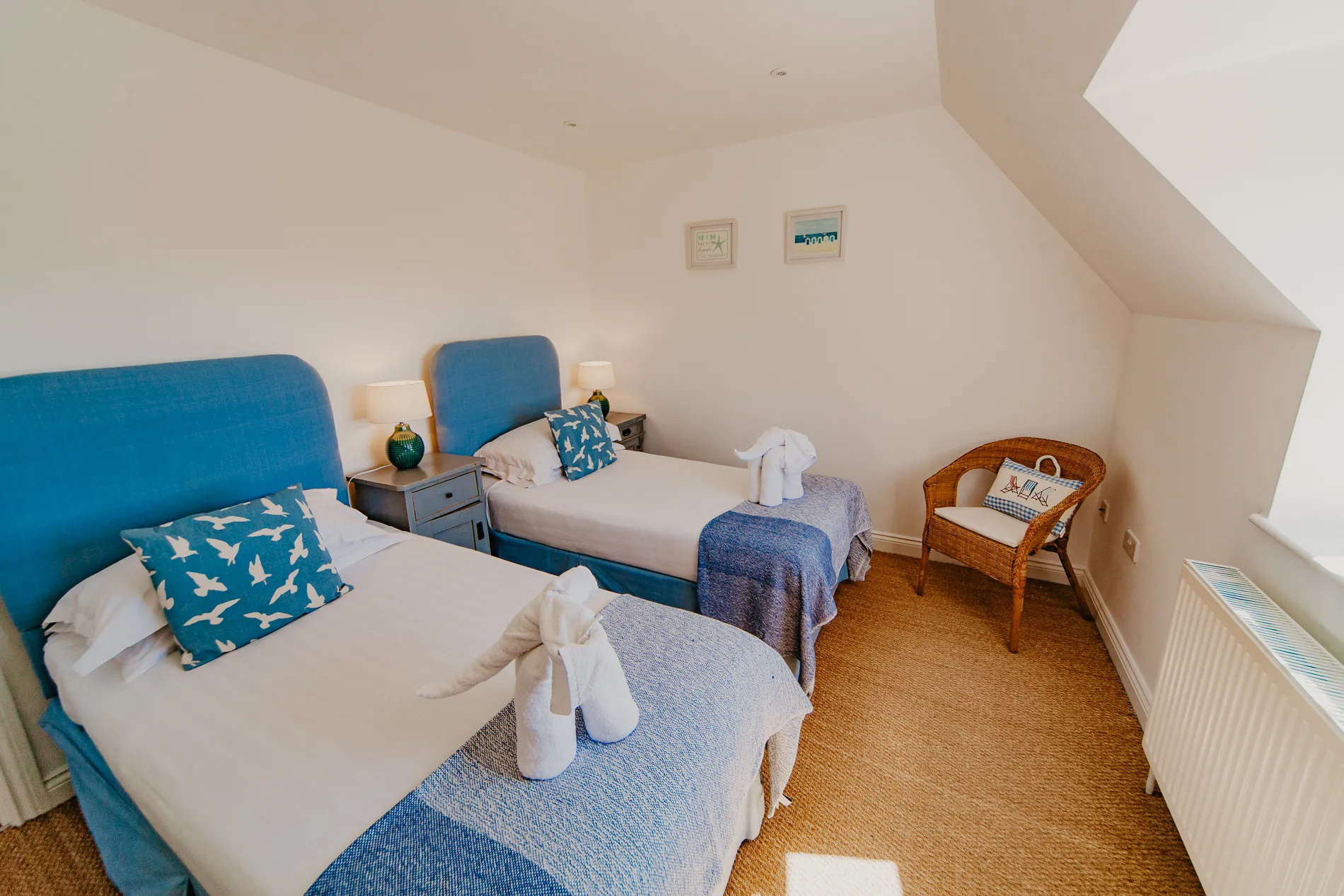 Twin bedroom with two single beds, blue headboards and throws, bedside lamps and a wicker chair under a sloping ceiling