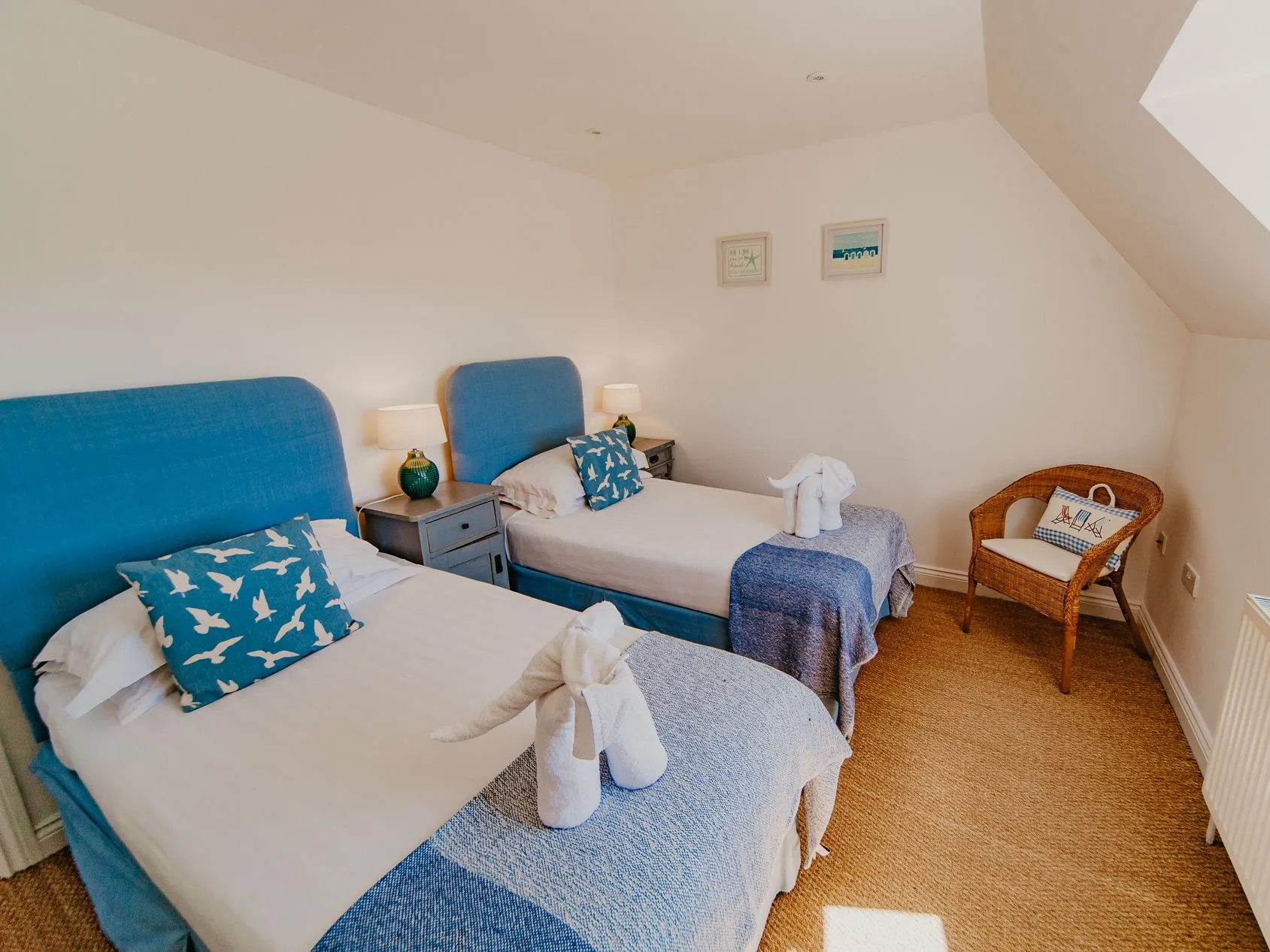 Twin bedroom with two single beds, blue headboards and throws, bedside lamps and a wicker chair under a sloping ceiling