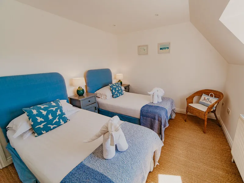 Twin bedroom with two single beds, blue headboards and throws, bedside lamps and a wicker chair under a sloping ceiling