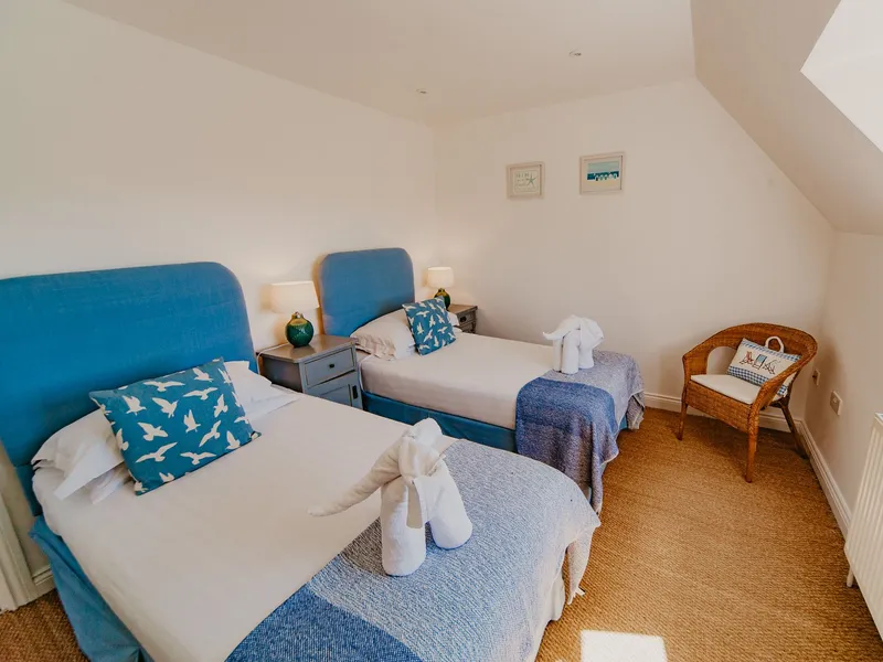 Twin bedroom with two single beds, blue headboards and throws, bedside lamps and a wicker chair under a sloping ceiling