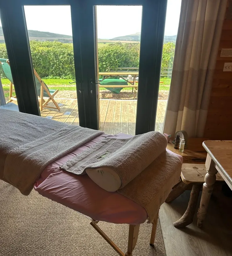 Massage table set up in a cosy wooden room overlooking a garden and rolling hills through glass doors