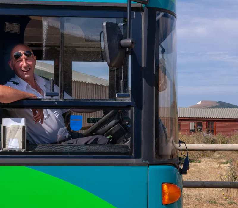 Smiling bus driver leaning out of the cab window of a turquoise bus on a sunny day.
