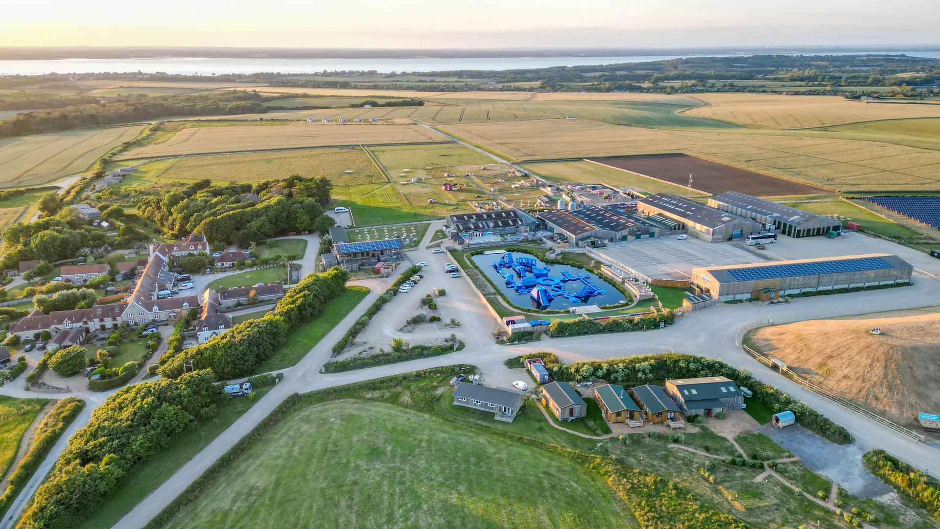 Drone view of Tapnell Farm with farm buildings, holiday accommodation, surrounding fields and coastline in the distance at sunset.