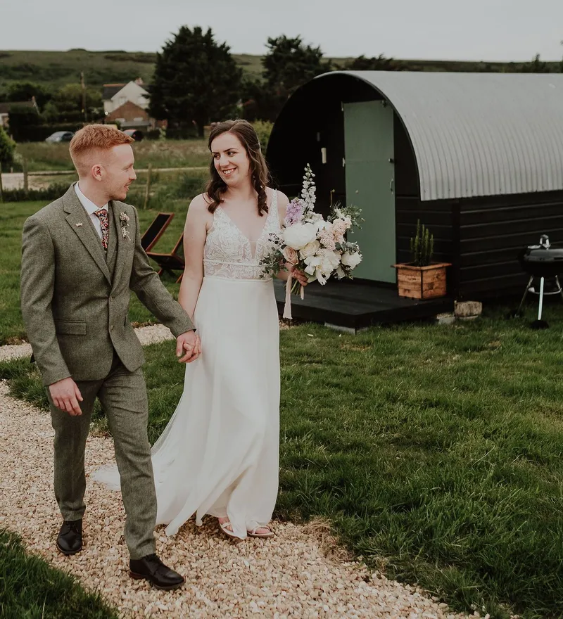 Bride and groom walking hand in hand on a gravel path beside a glamping pod in a countryside setting