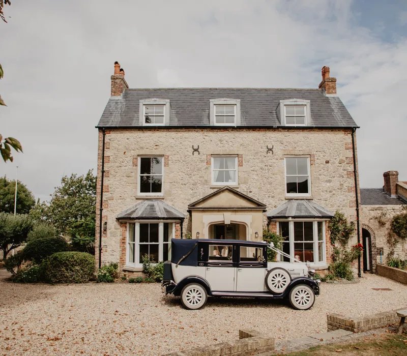 Vintage wedding car parked outside a large stone country house with a gravel driveway.