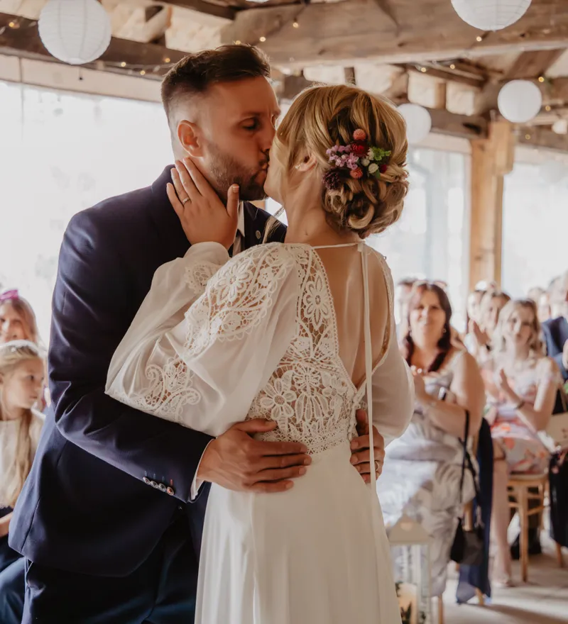 Bride and groom sharing a kiss during a rustic indoor wedding ceremony as guests look on