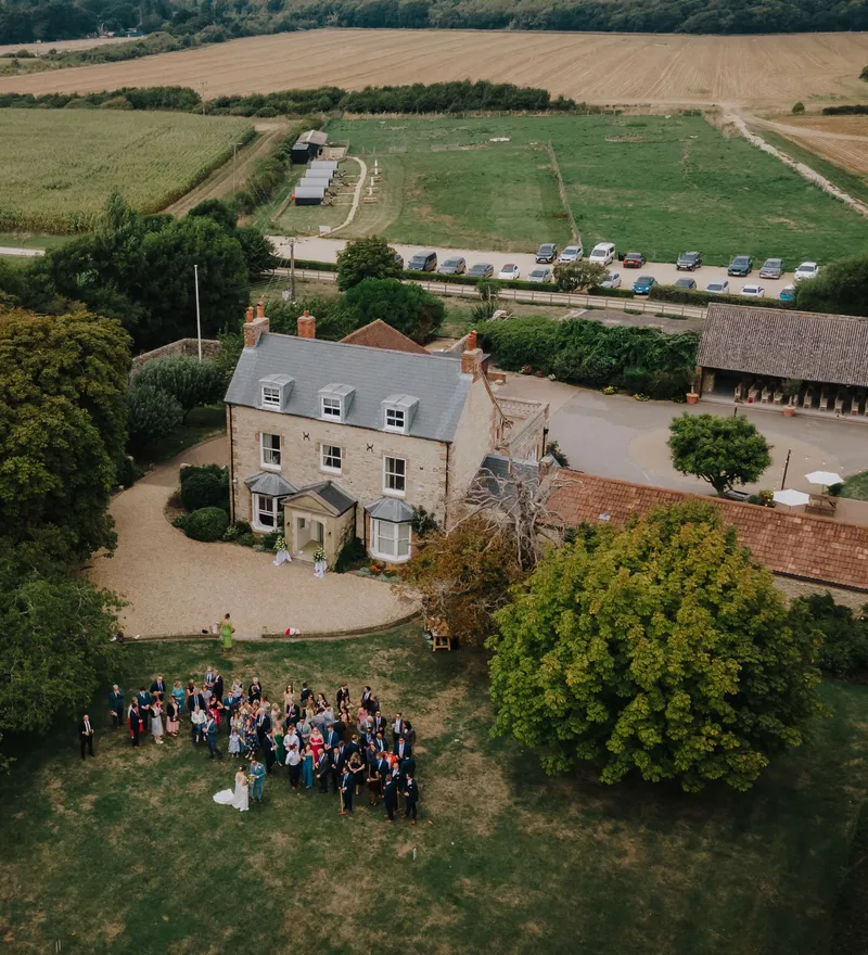 Aerial view of wedding guests gathered on the lawn outside a countryside manor house surrounded by fields and trees.